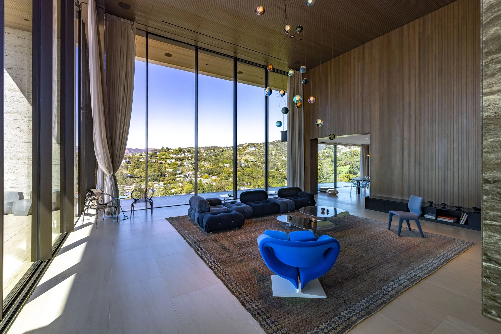 Modern living room with large floor-to-ceiling windows showcasing scenic hillside view, contemporary blue and black seating, glass coffee tables on a patterned rug, high wooden ceiling with hanging globe light fixtures, and minimalist decor.