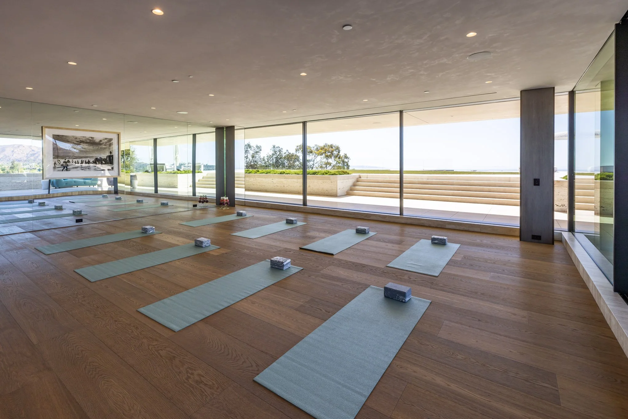 Sunlit yoga studio with wooden floors, gray yoga mats and blocks arranged for a class, large windows with a view of stairs and outdoor greenery.