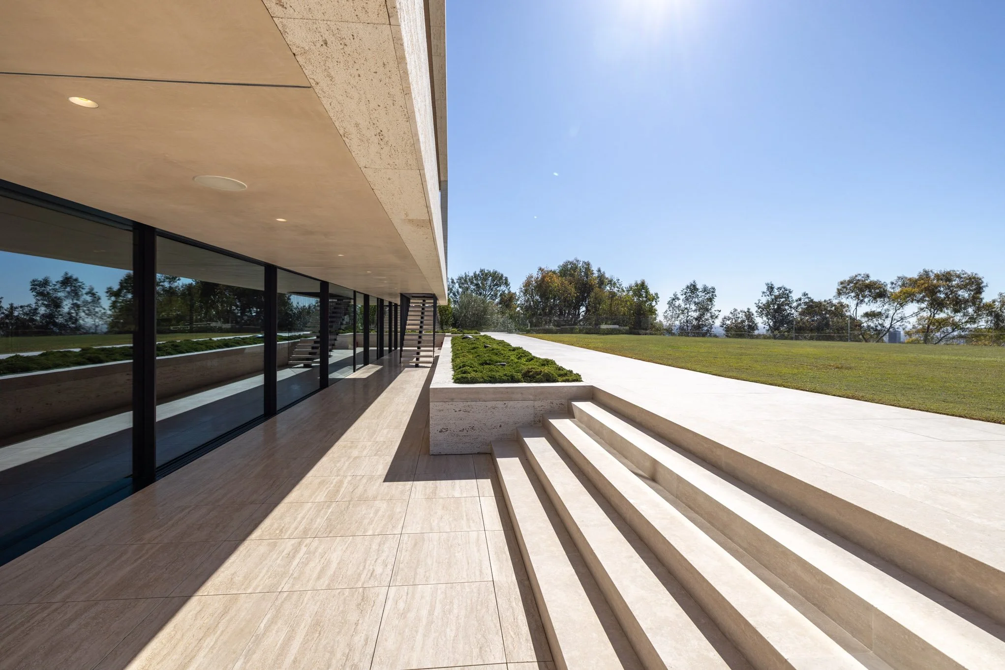 Modern building with large glass windows, beige tiled floor, and steps leading to an outdoor grassy area under a clear blue sky.