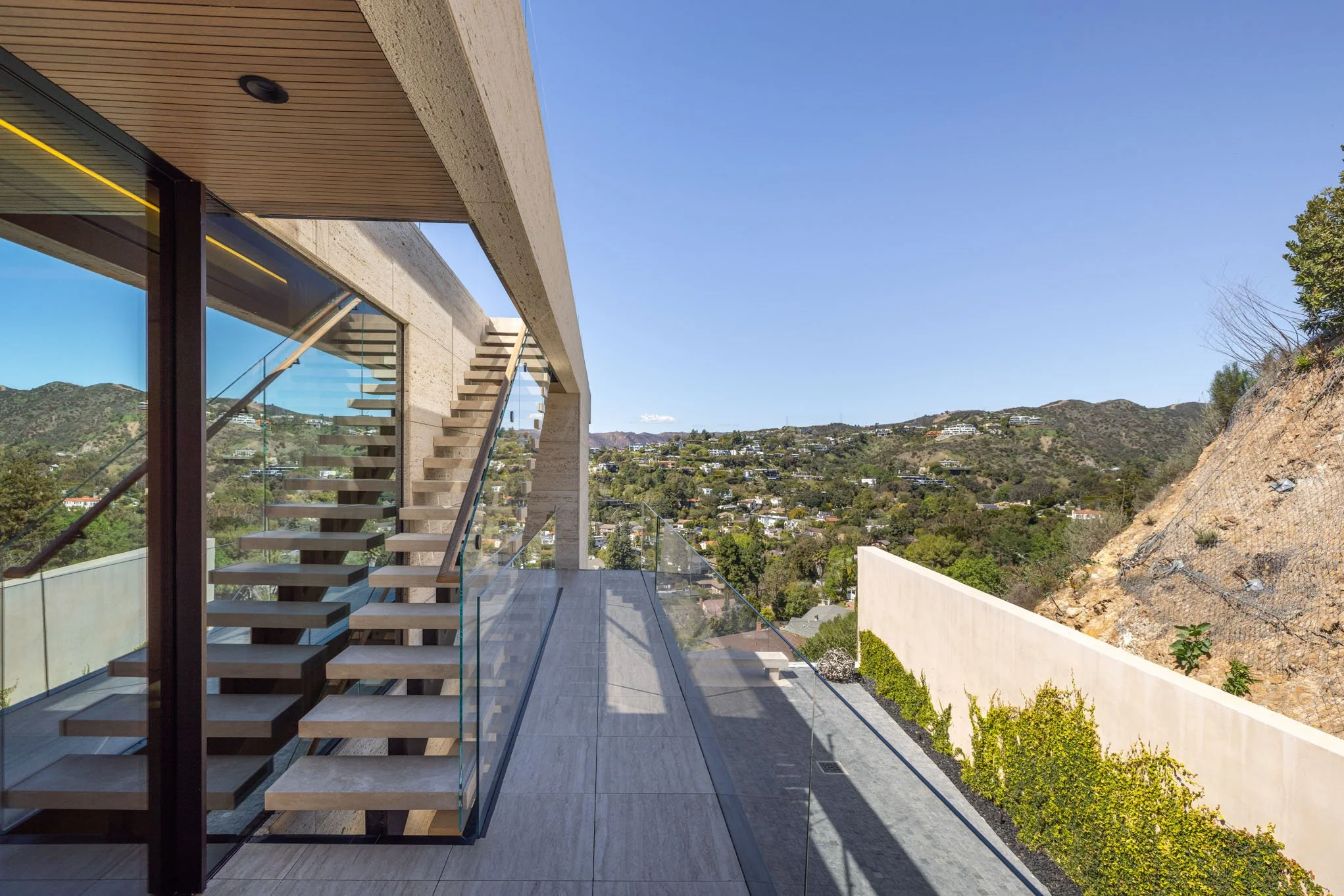 Modern outdoor balcony with glass railing, overlooking a hilly landscape and clear blue sky, featuring a staircase visible inside through large glass windows.