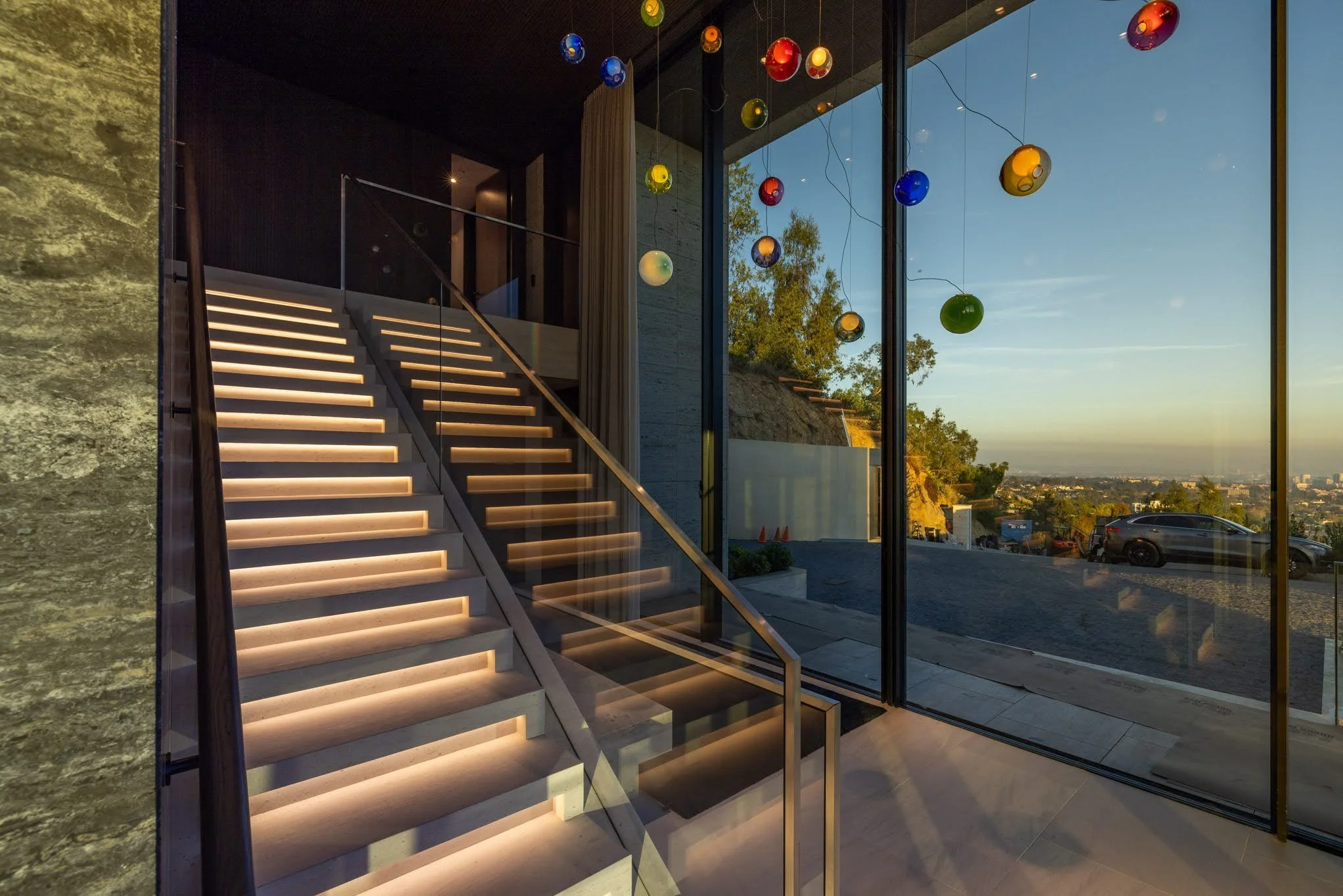 Modern indoor staircase with LED lighting, large glass windows, and colorful hanging glass orbs, overlooking a scenic outdoor view during sunset.