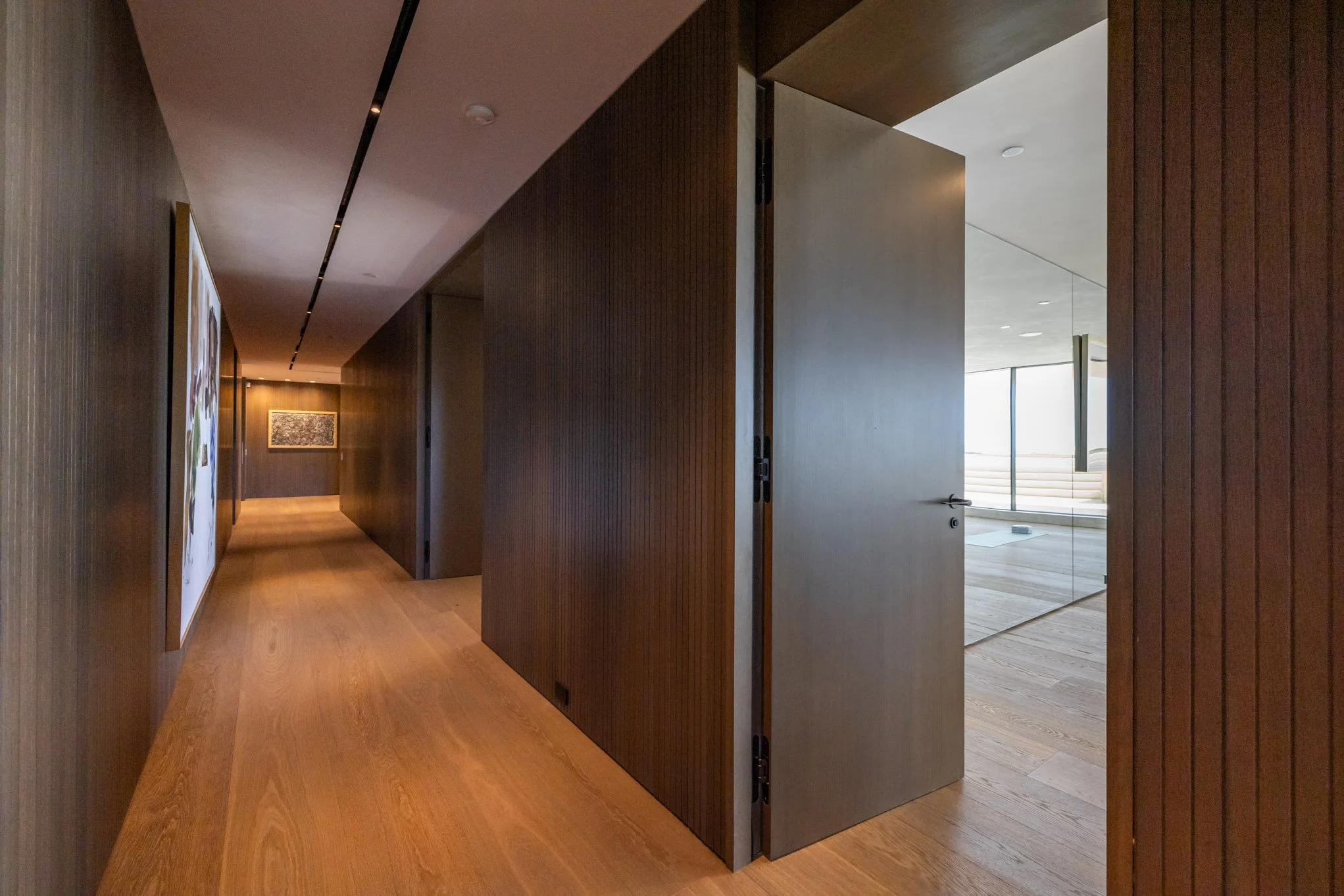 Modern interior hallway with wood-paneled walls, wooden flooring, and an open door leading to a bright room with large windows and a view of steps outside.