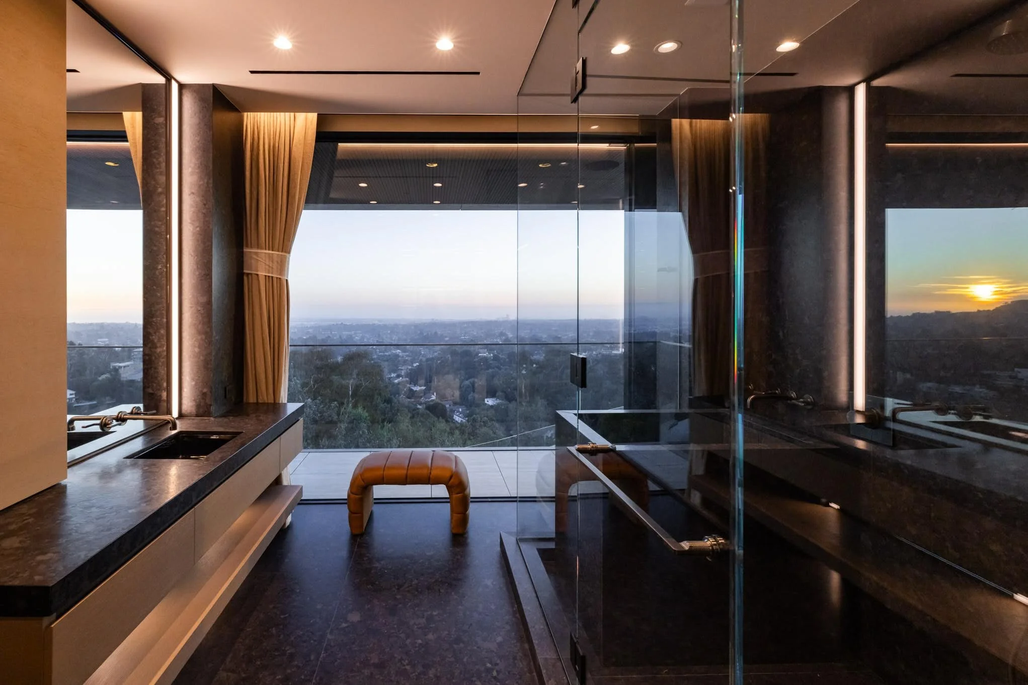 Modern bathroom with large backlit mirror, black countertops, a small stool, and a glass-enclosed shower, overlooking a city view at sunset.