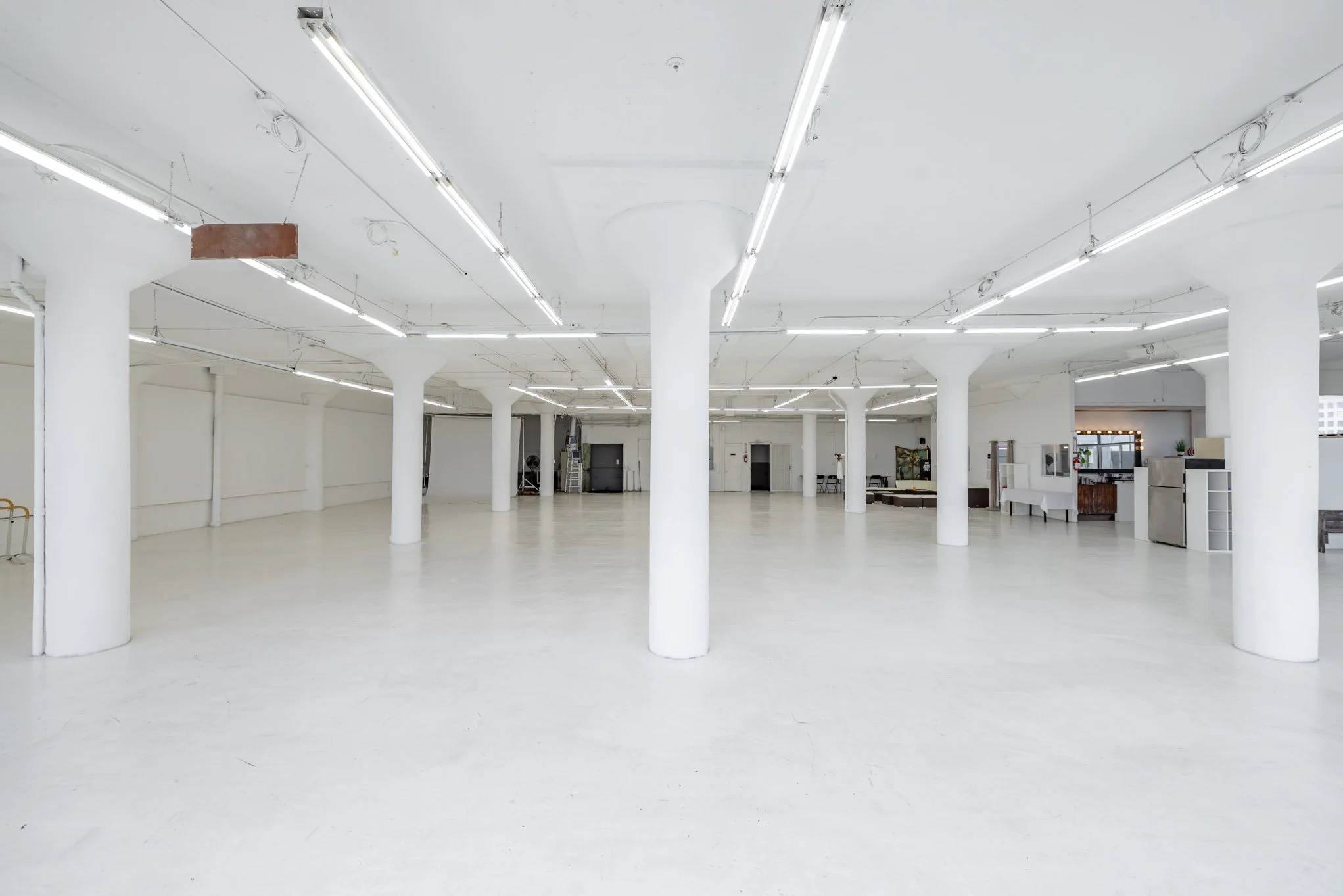 Large, empty, white-walled indoor space with multiple white columns and fluorescent lighting fixtures.