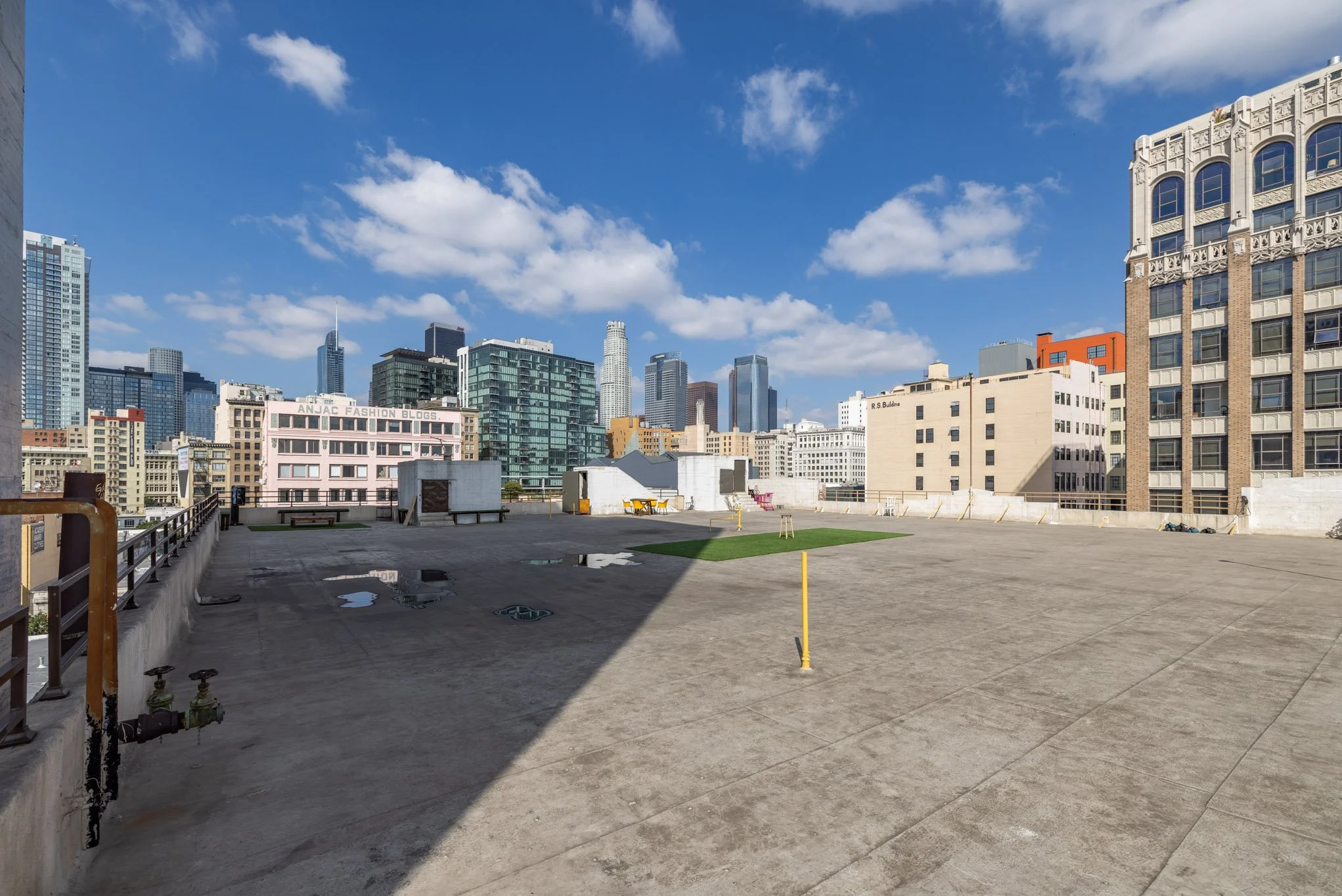 Empty rooftop with concrete surface, small patches of grass, and city skyline with modern high-rise buildings under a blue sky with clouds.