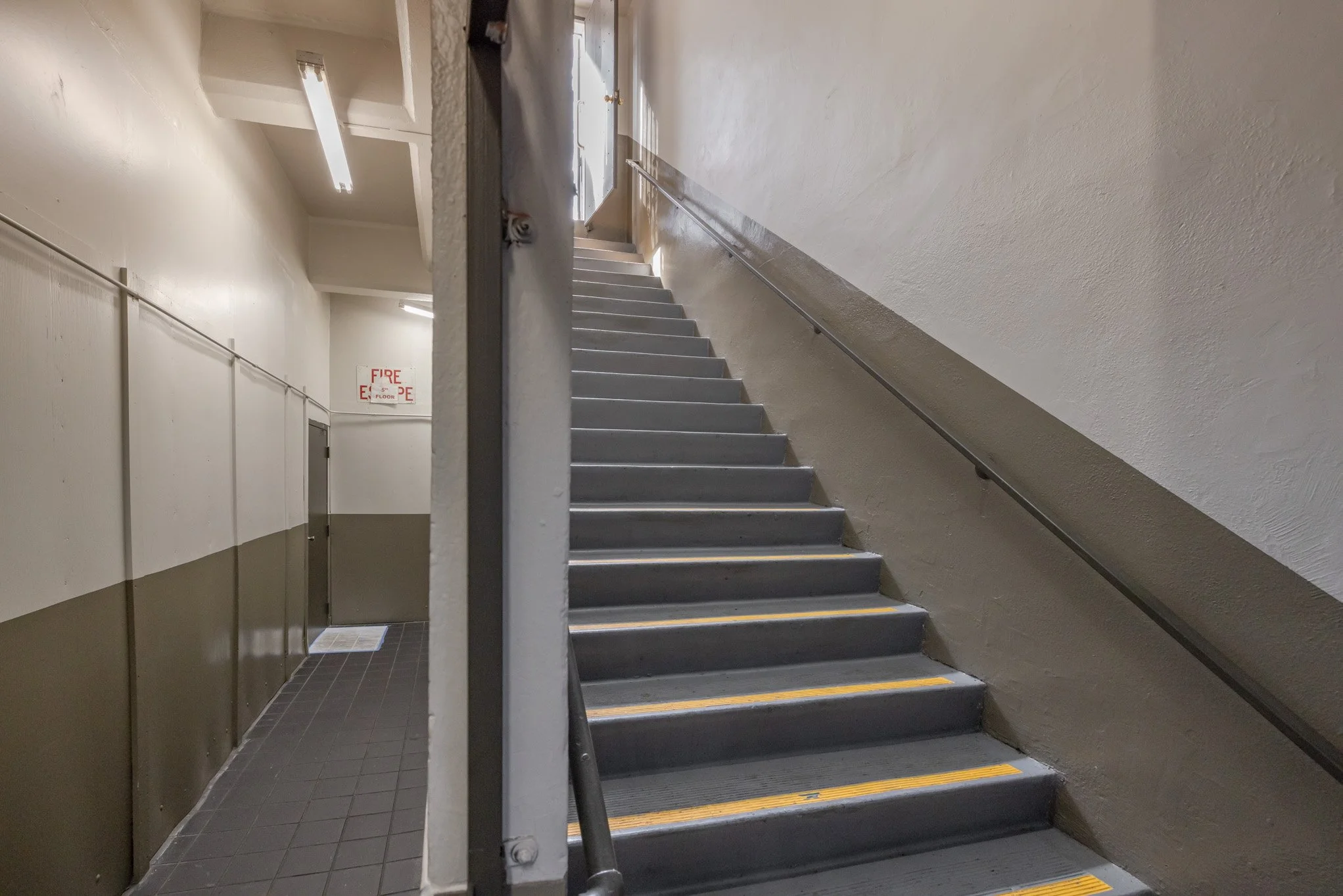 A stairway inside a building with a metal handrail on the right side, leading upwards, with a partial view of another staircase or platform at the top. The walls are painted beige and the stairs are gray with yellow markings on the edges. There is a 