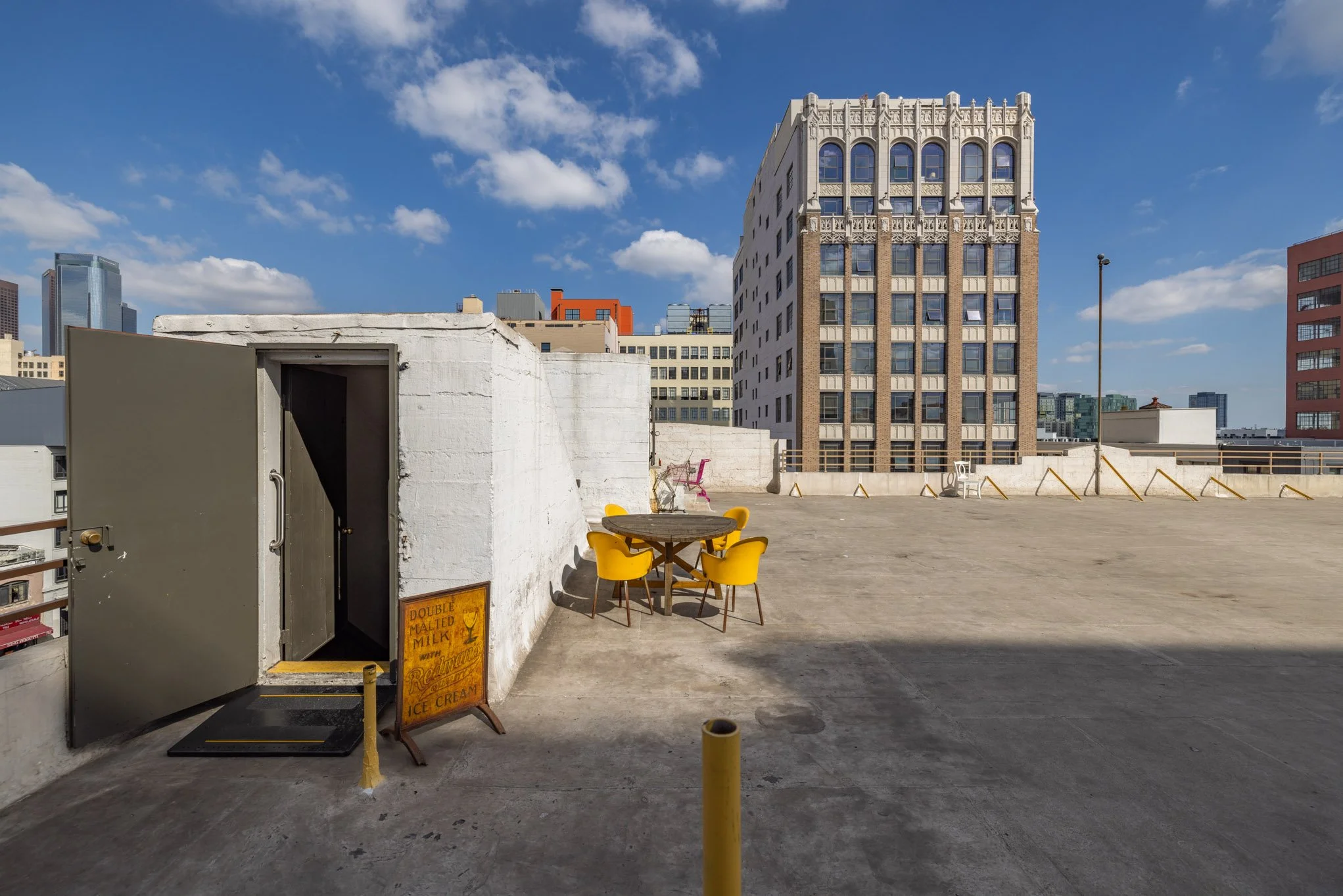 A rooftop with a round table and four yellow chairs, a pink chair, and a white chair. There is a white wall with an open door and a sign advertising double malted milk with Rayben ice cream. The background shows a city skyline with various buildings 