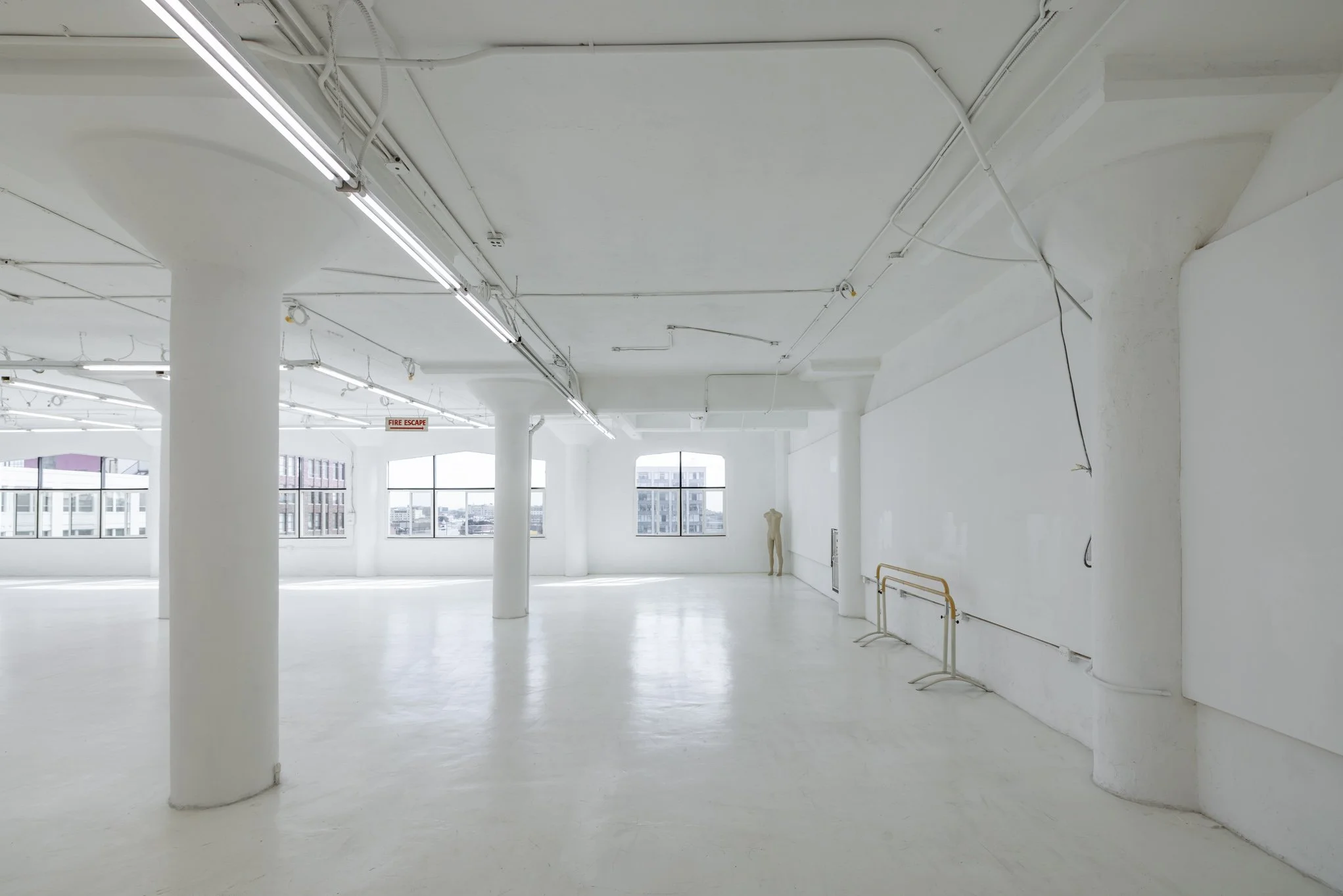 Empty white interior space with large windows, white columns, ceiling pipes, and a fire escape sign.