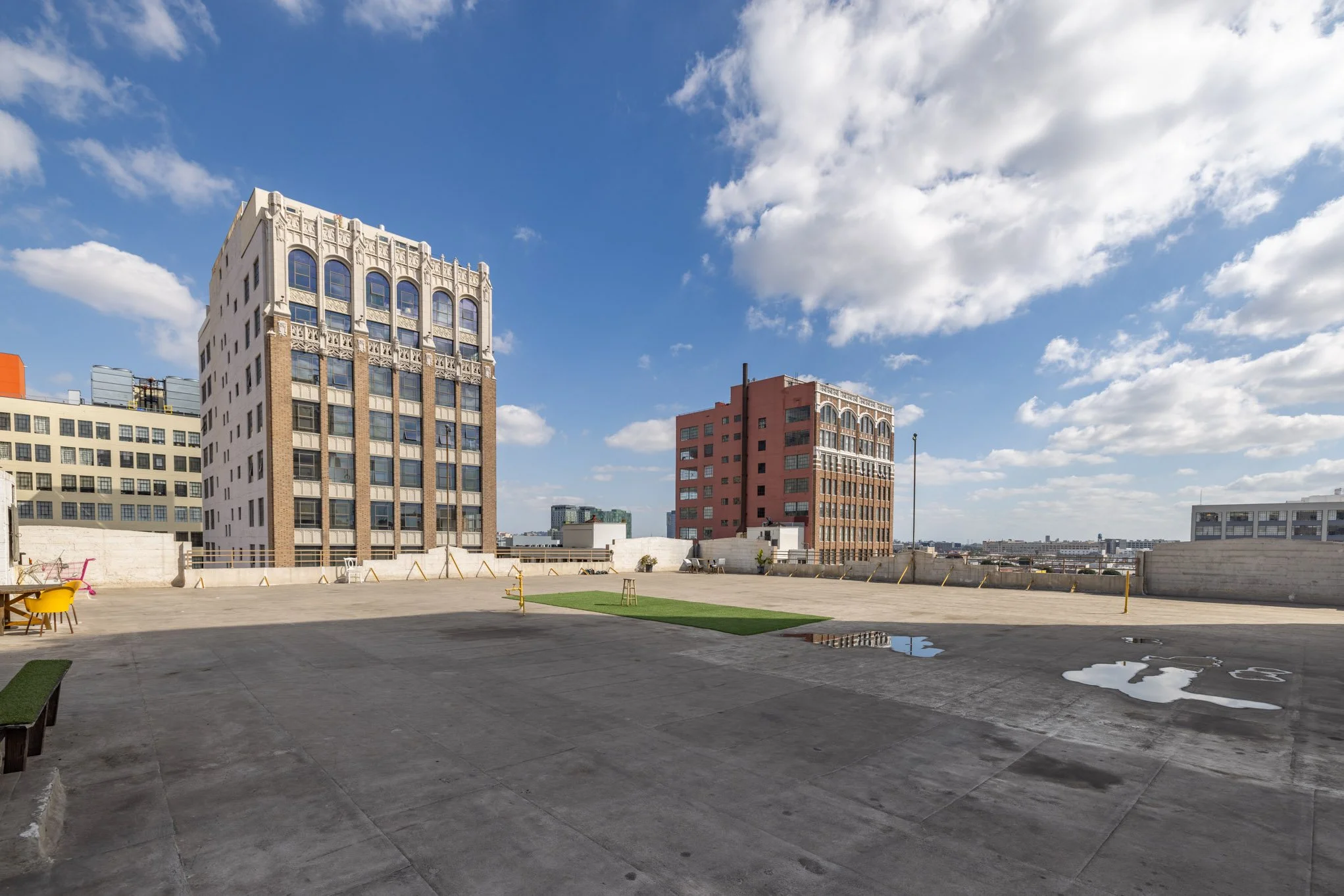 Cloudy sky over a rooftop parking lot with puddles, some yellow chairs, and tall buildings in the distance.
