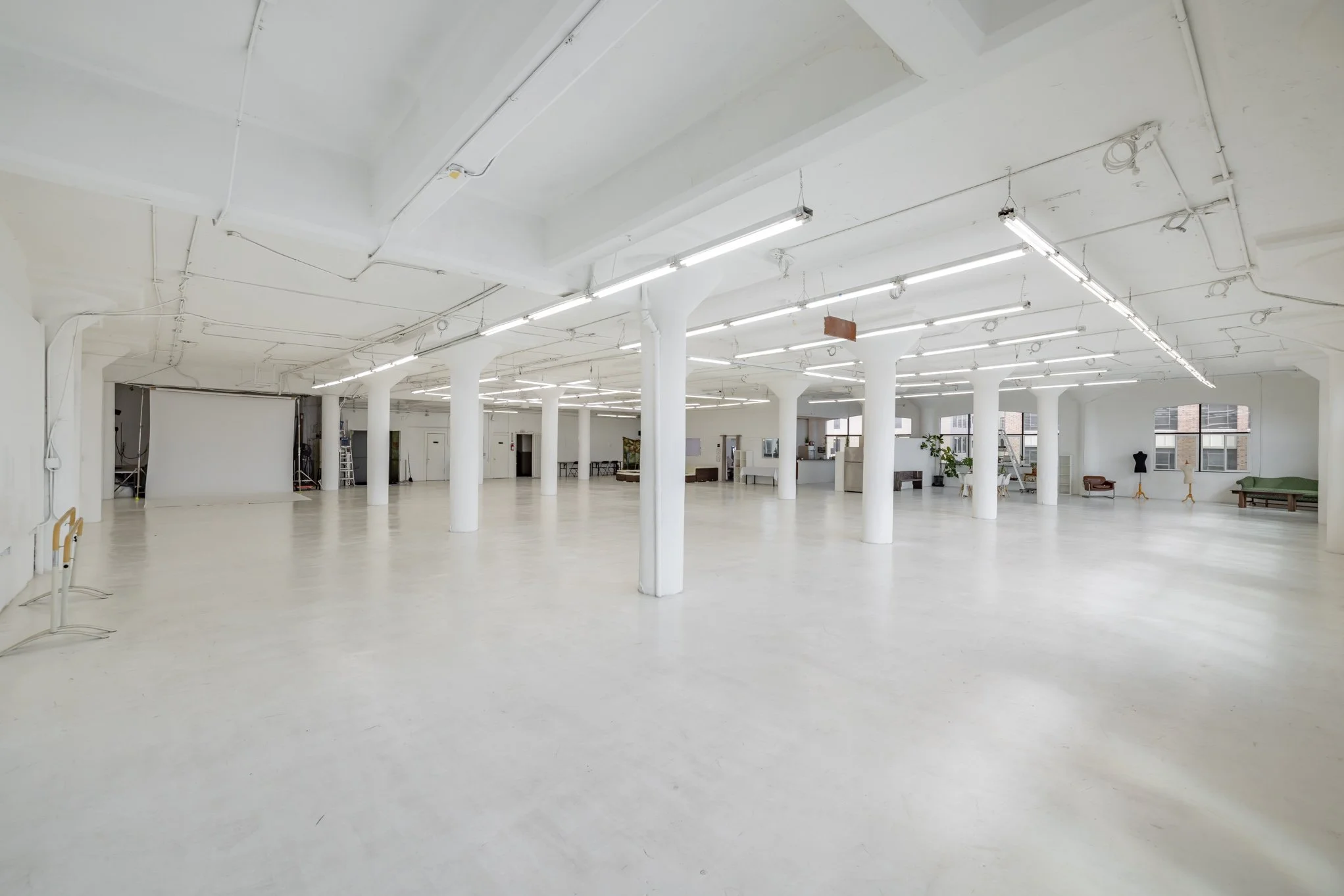 A spacious, empty, white studio space with high ceilings, ceiling lights, columns, large windows, and some furniture against the walls.