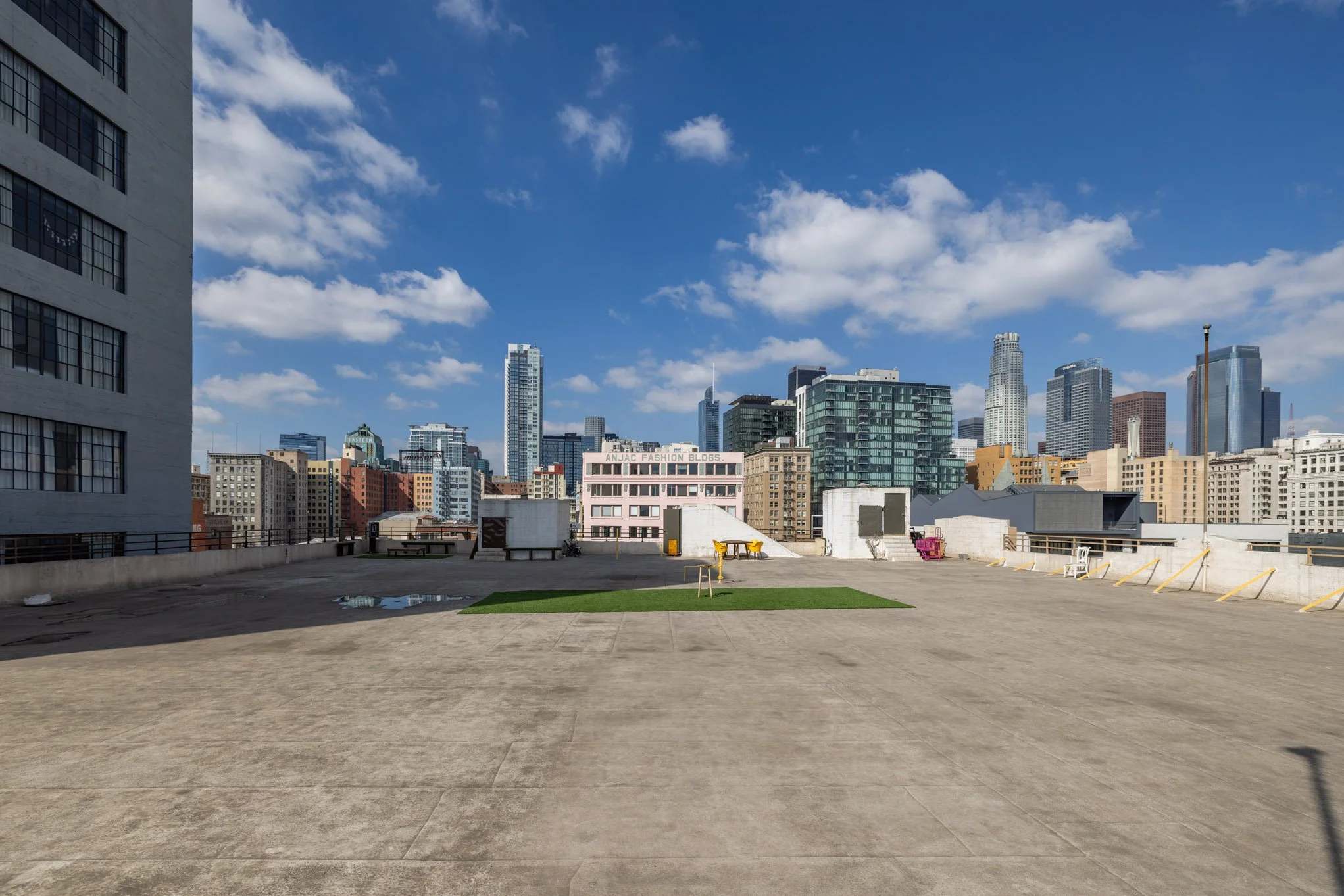 Empty rooftop parking area in a city with tall buildings under a blue sky with scattered clouds.