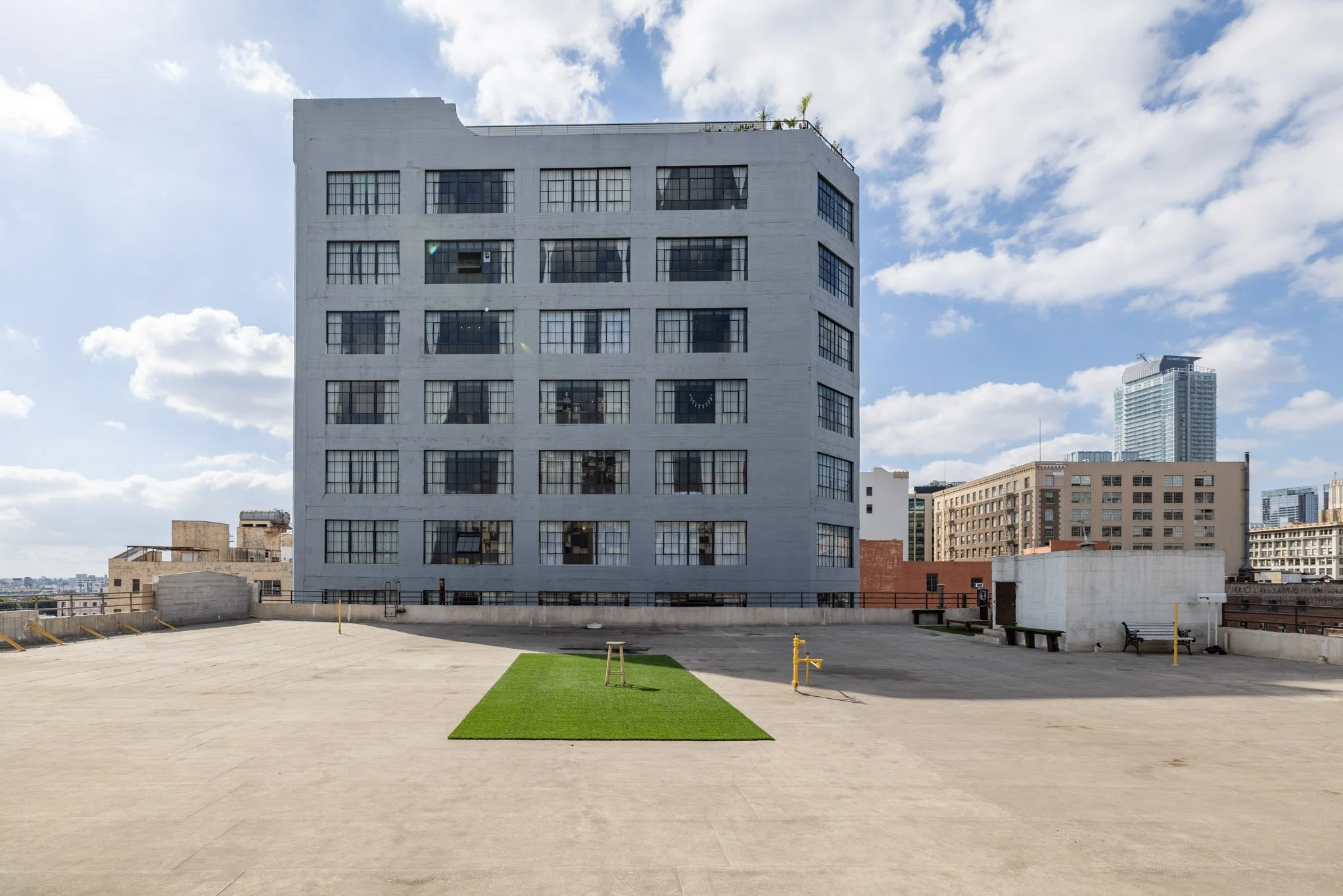 City rooftop with a large gray building, a small patch of artificial grass, and surrounding cityscape with taller buildings in the background.