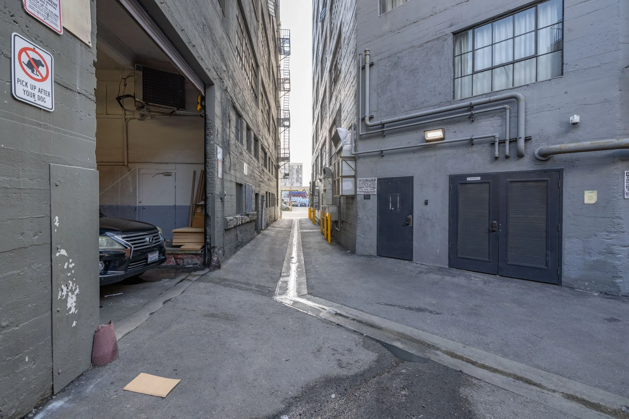 Urban alleyway with a parking garage and a car partially visible on the left, gray factory-style walls with pipes, doors, and signs, and a narrow alley leading to a distant parking lot.
