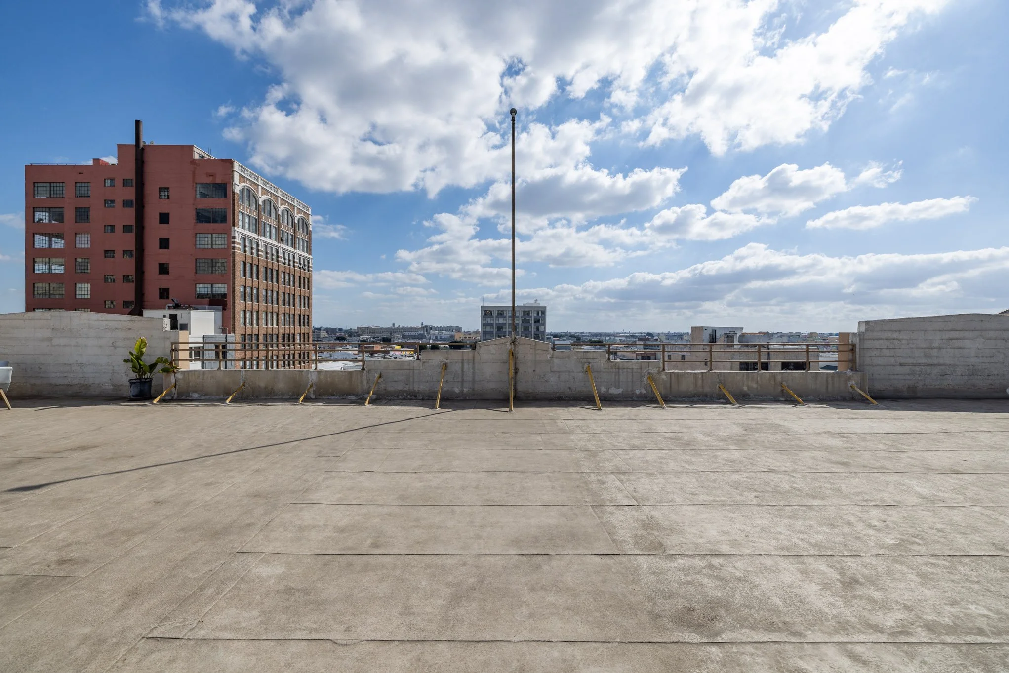 City rooftop parking area with concrete floor, metal railings, a potted plant, and a large building with red and beige brick exterior in the background under a partly cloudy sky.