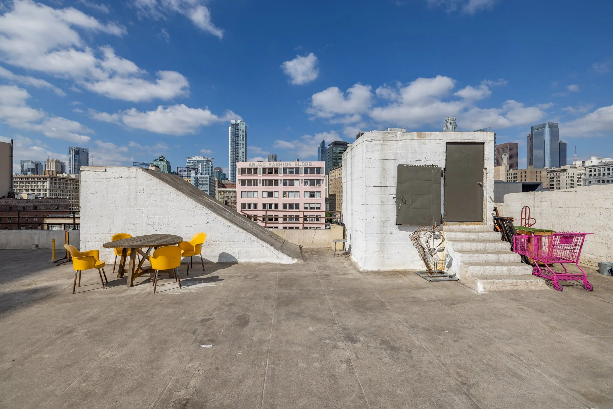 Rooftop terrace in a city with a round table and four yellow chairs, pink shopping cart, and city skyline in the background under a blue sky with scattered clouds.