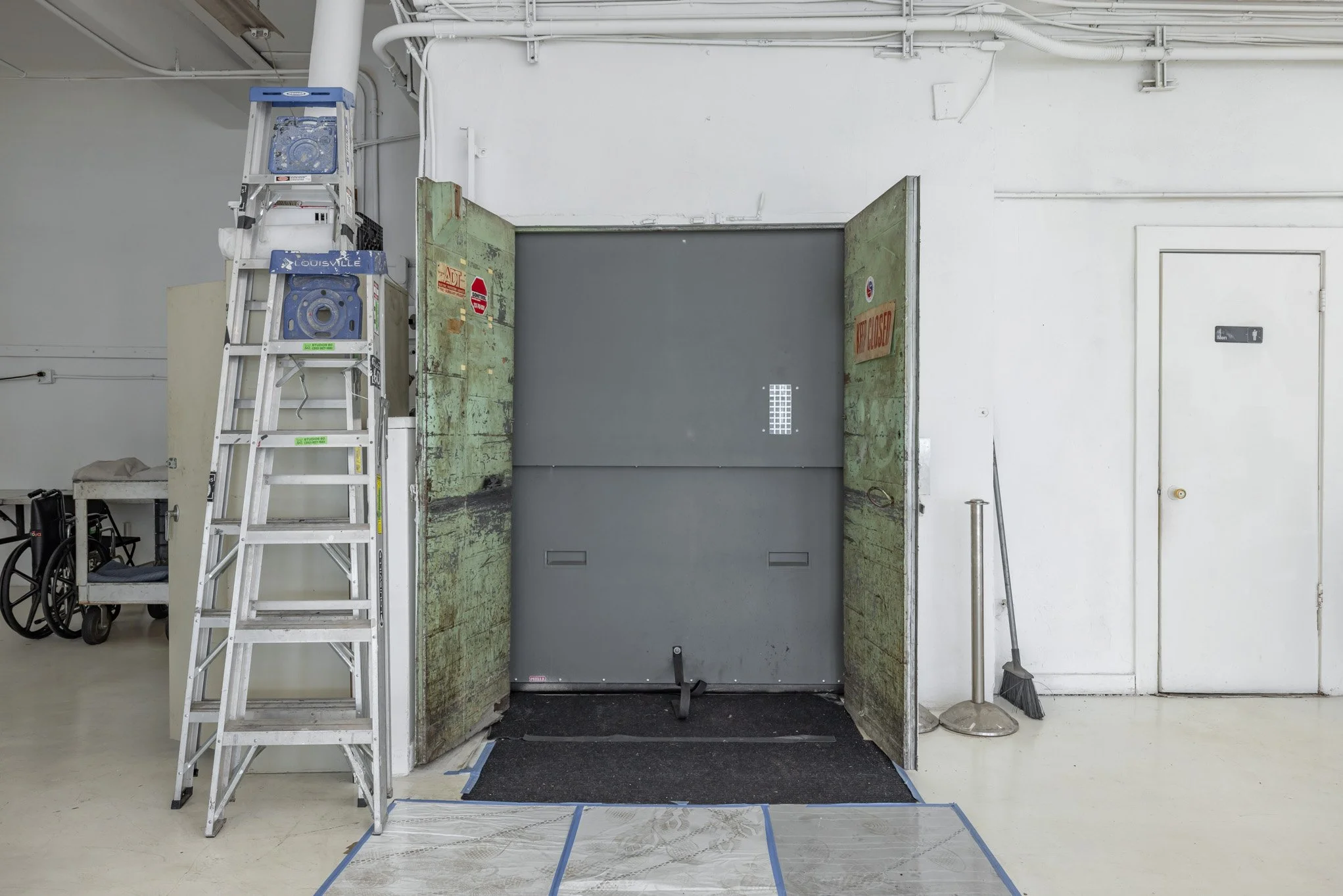 A large metal door in an industrial or utility area, partially open, with cleaning tools and a ladder nearby.