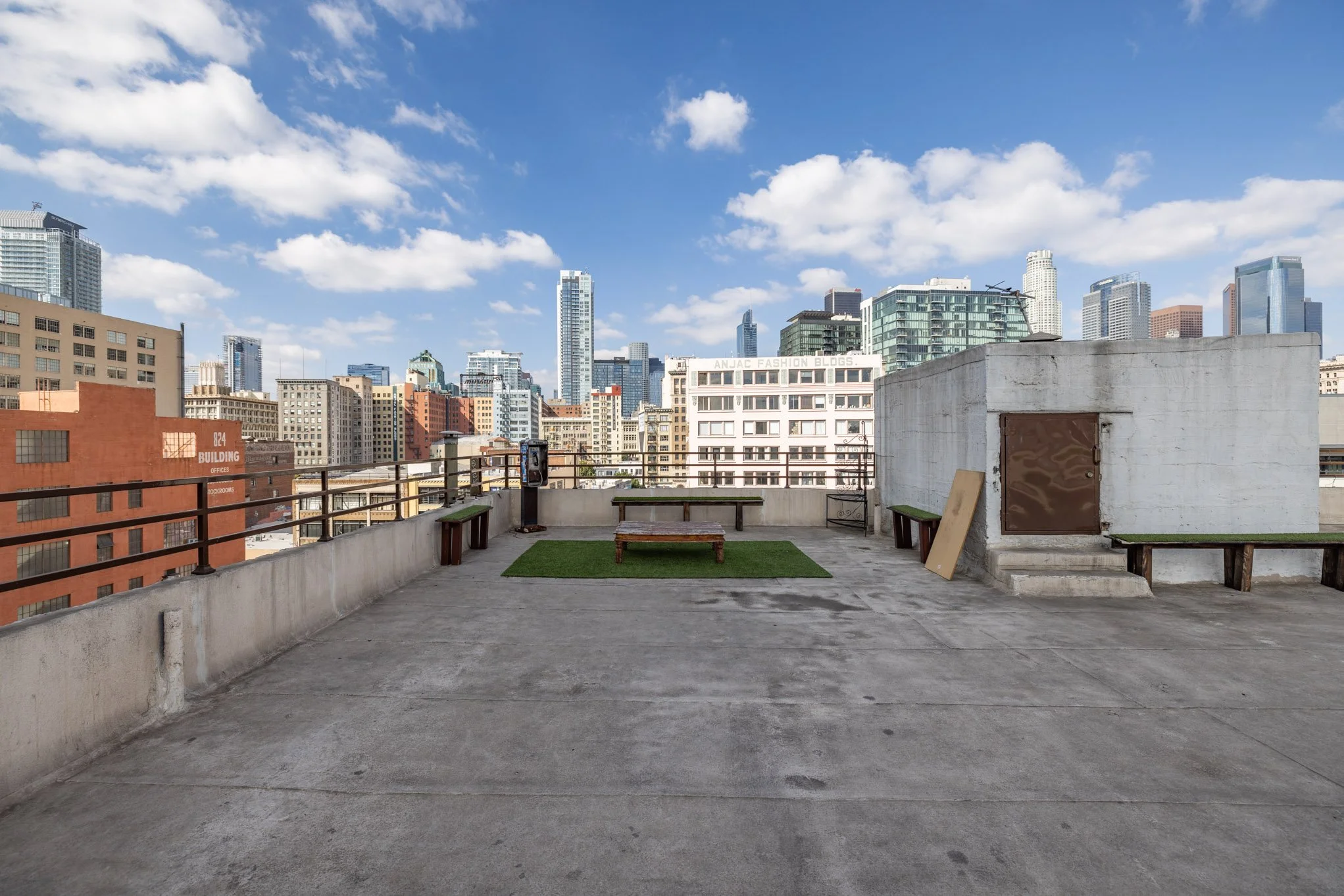 Rooftop view of a city with tall buildings, benches, a small patch of grass, and a clear blue sky with scattered clouds.
