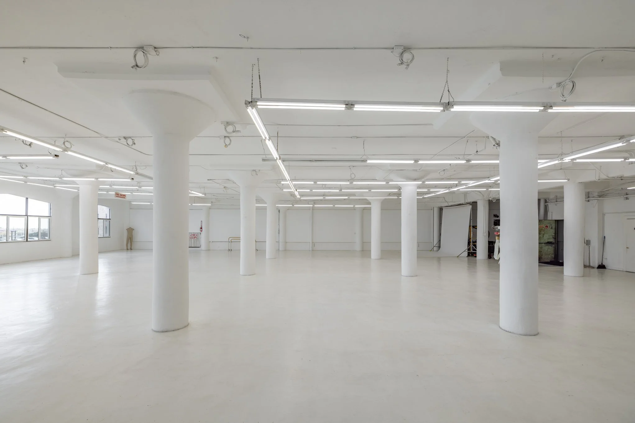 Empty, white-walled industrial space with large support columns, fluorescent lighting, and windows.
