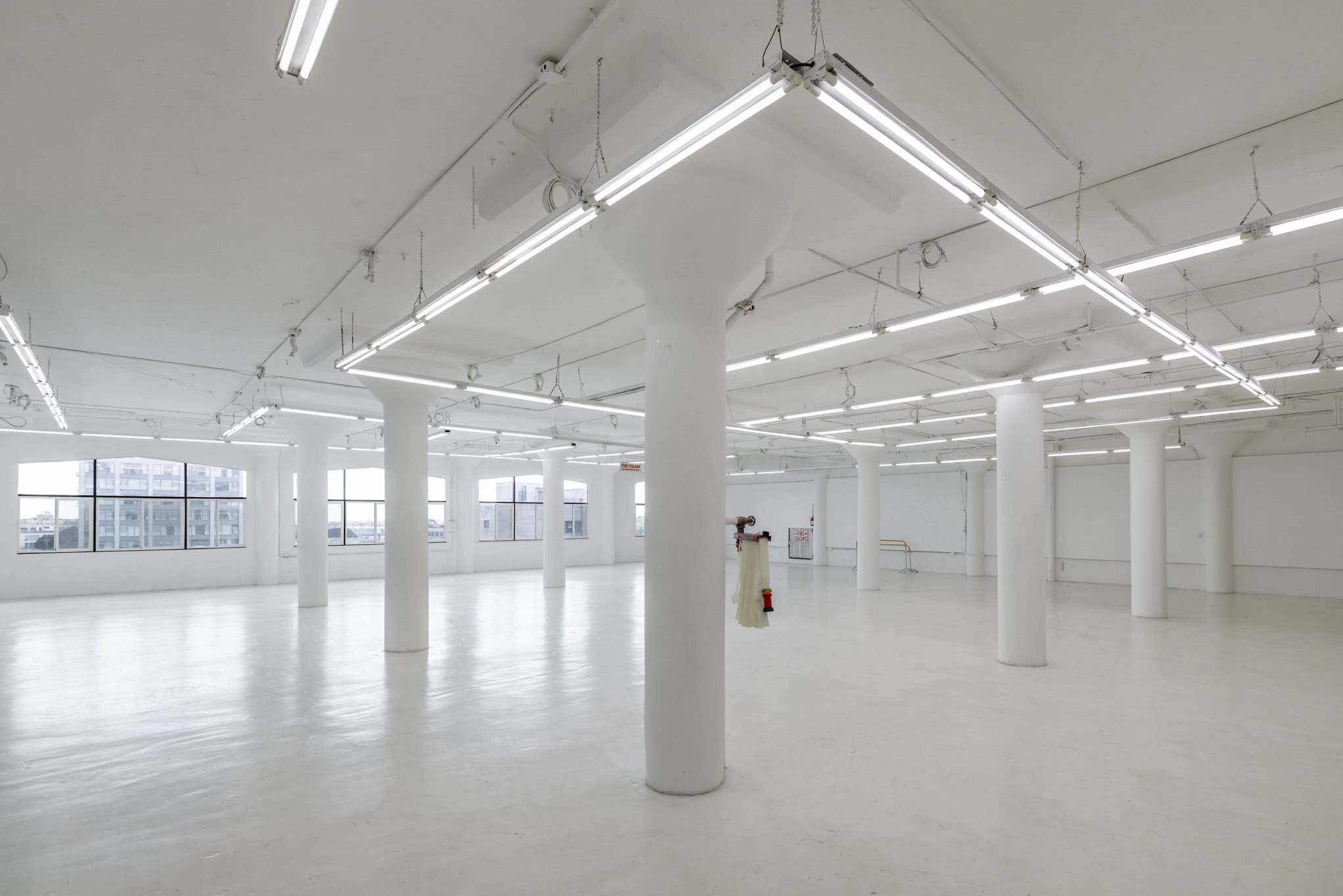 Empty, white-walled industrial or commercial space with large windows, white support columns, and bright fluorescent lighting fixtures.