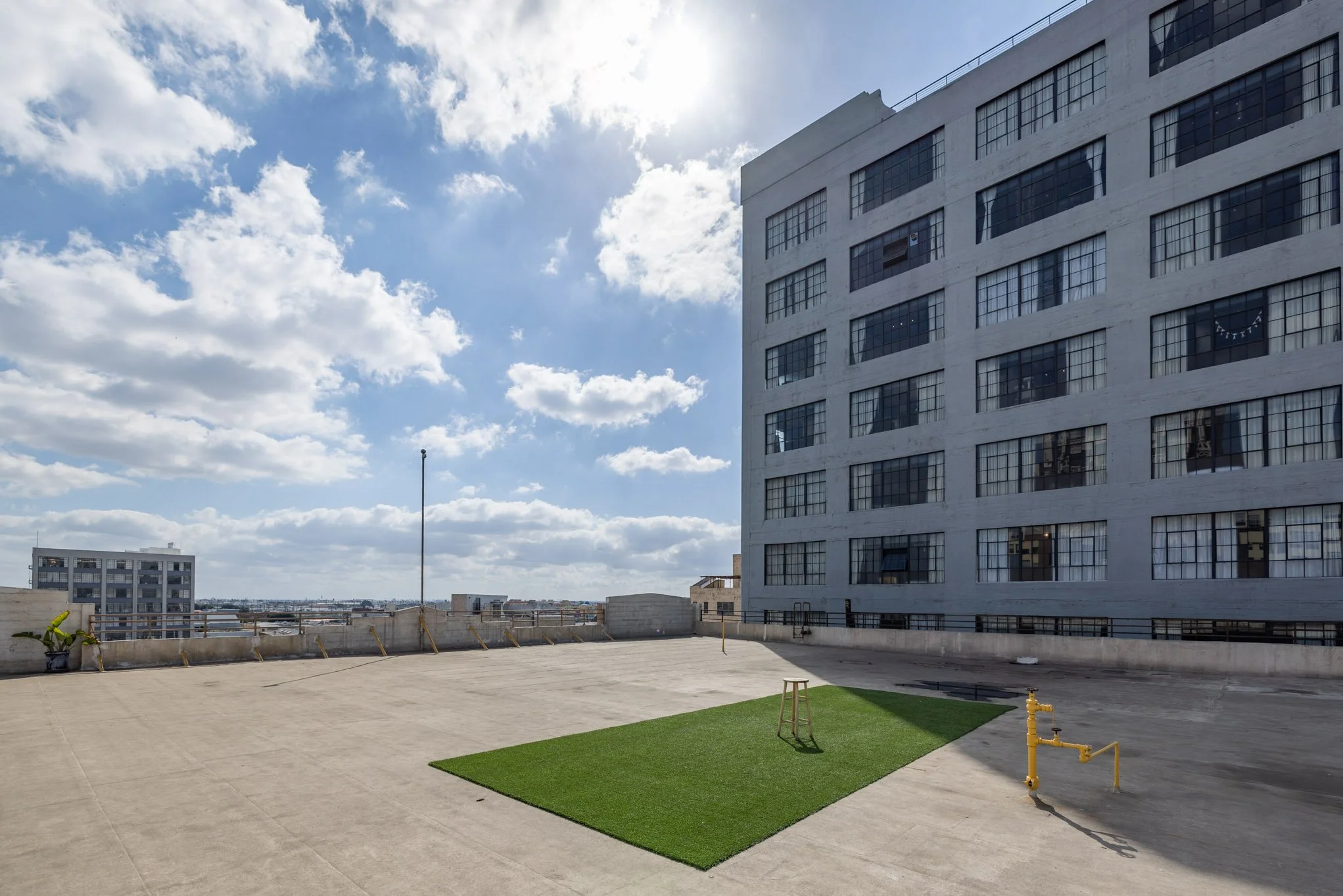 Empty rooftop with green turf, a small stool, and a fire hydrant, surrounded by concrete edges, under a partly cloudy sky with high-rise buildings in the background.