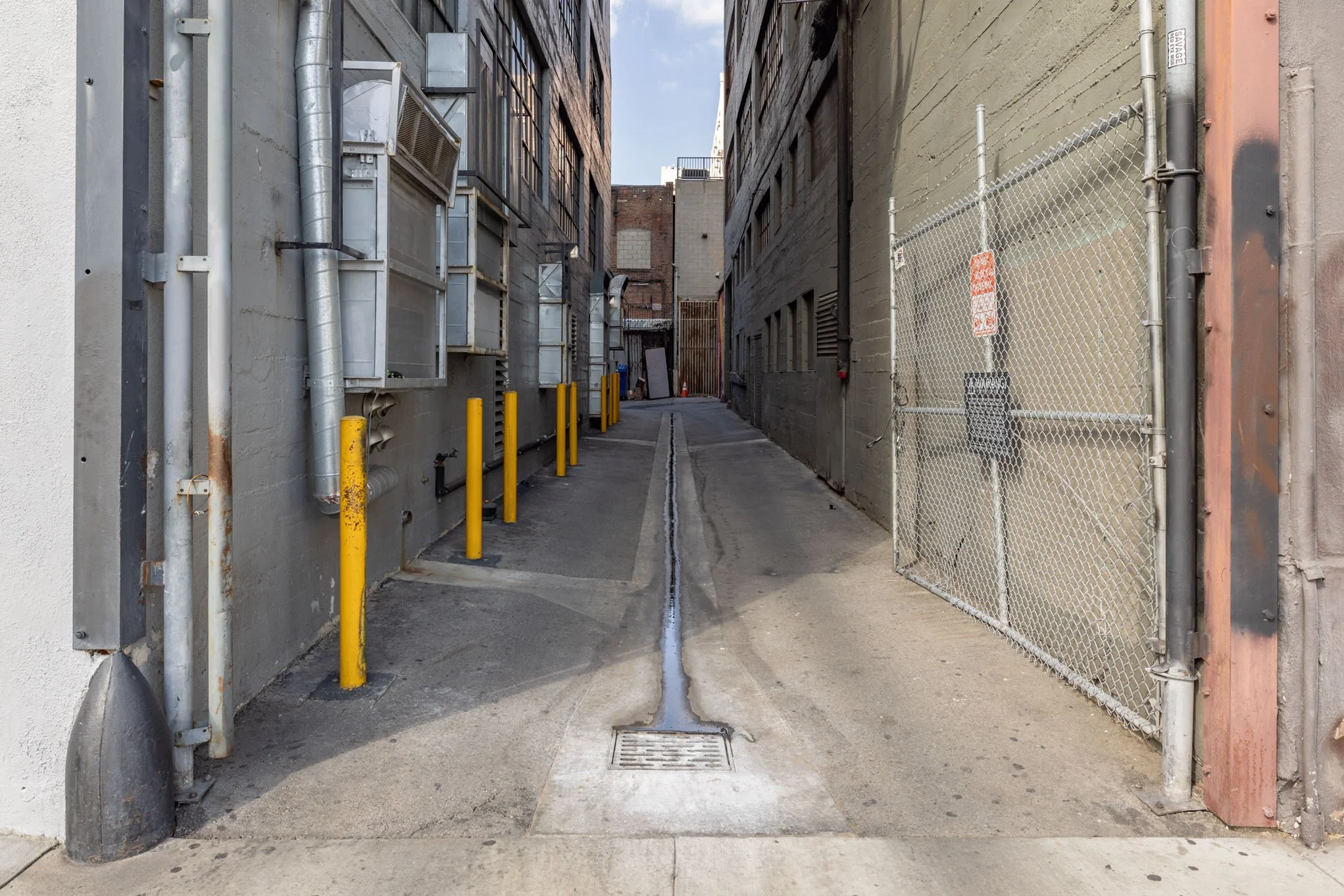 An alleyway between two buildings with metal pipes, yellow bollards, a drain in the ground, and a chain-link gate on the right side.