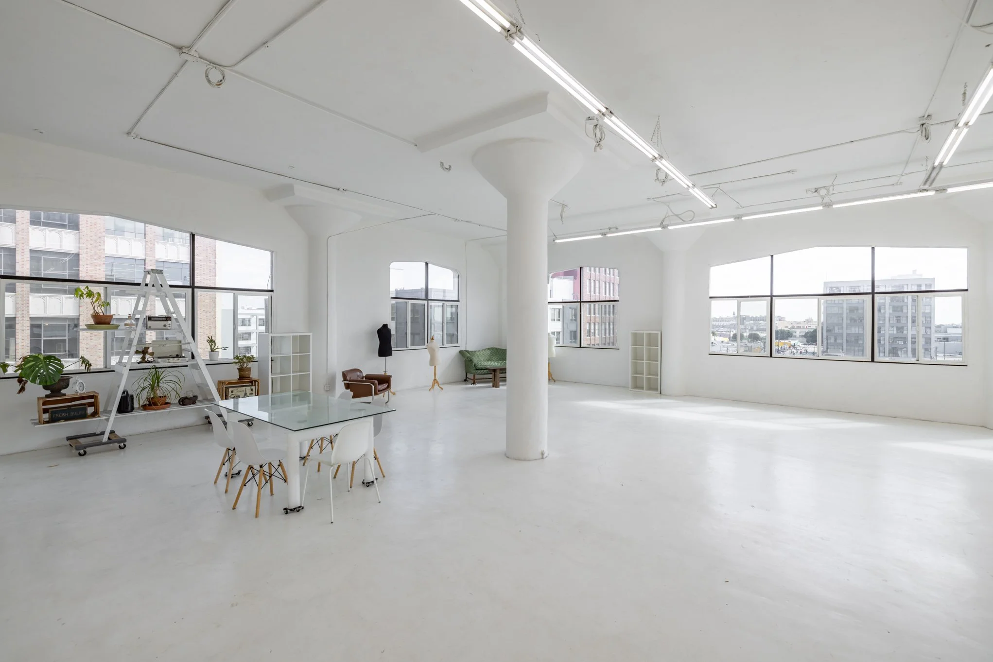 Interior of a spacious, bright, white art studio or gallery with large windows, a glass table with white chairs, shelves, and mannequins.