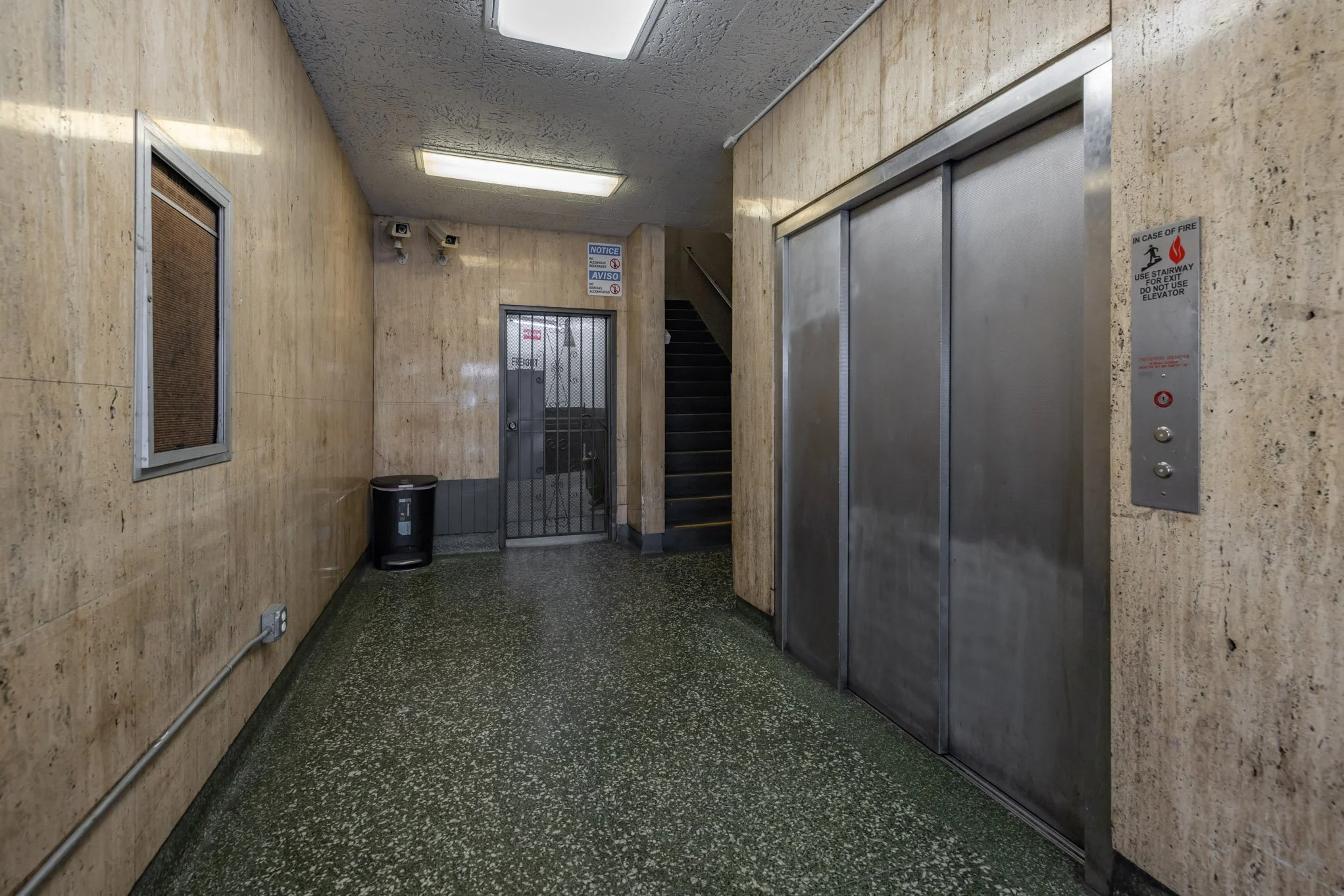 Indoor hallway with elevator, staircase, and door, beige walls, green speckled floor, ceiling lights, and security cameras.