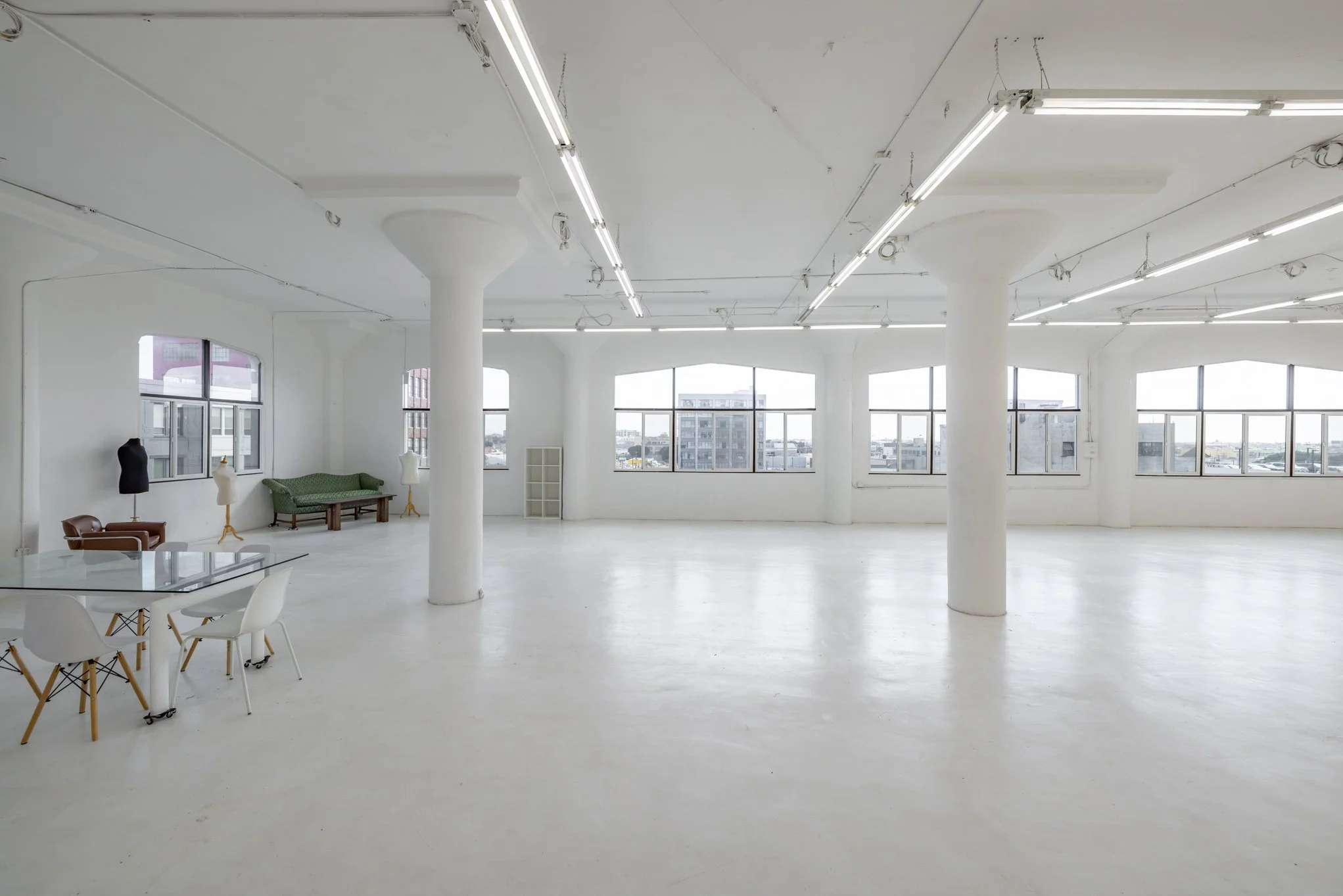 Spacious, empty white room with large windows, multiple white pillars, and minimal furniture including chairs, sofas, dress forms, and a photo table, illuminated by ceiling fluorescent lights.
