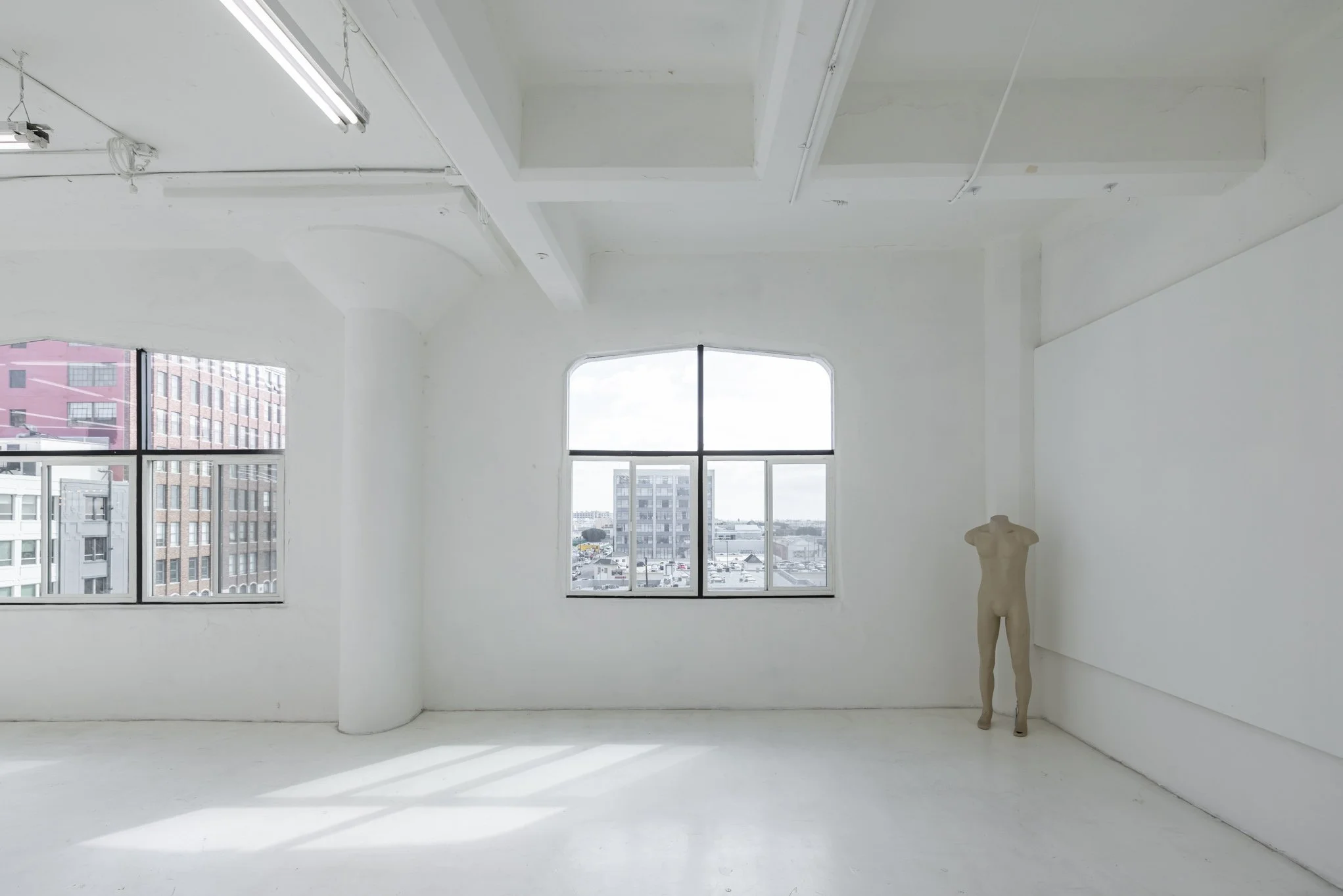 Empty white room with large windows, a torso mannequin standing in the corner, and city buildings visible outside.