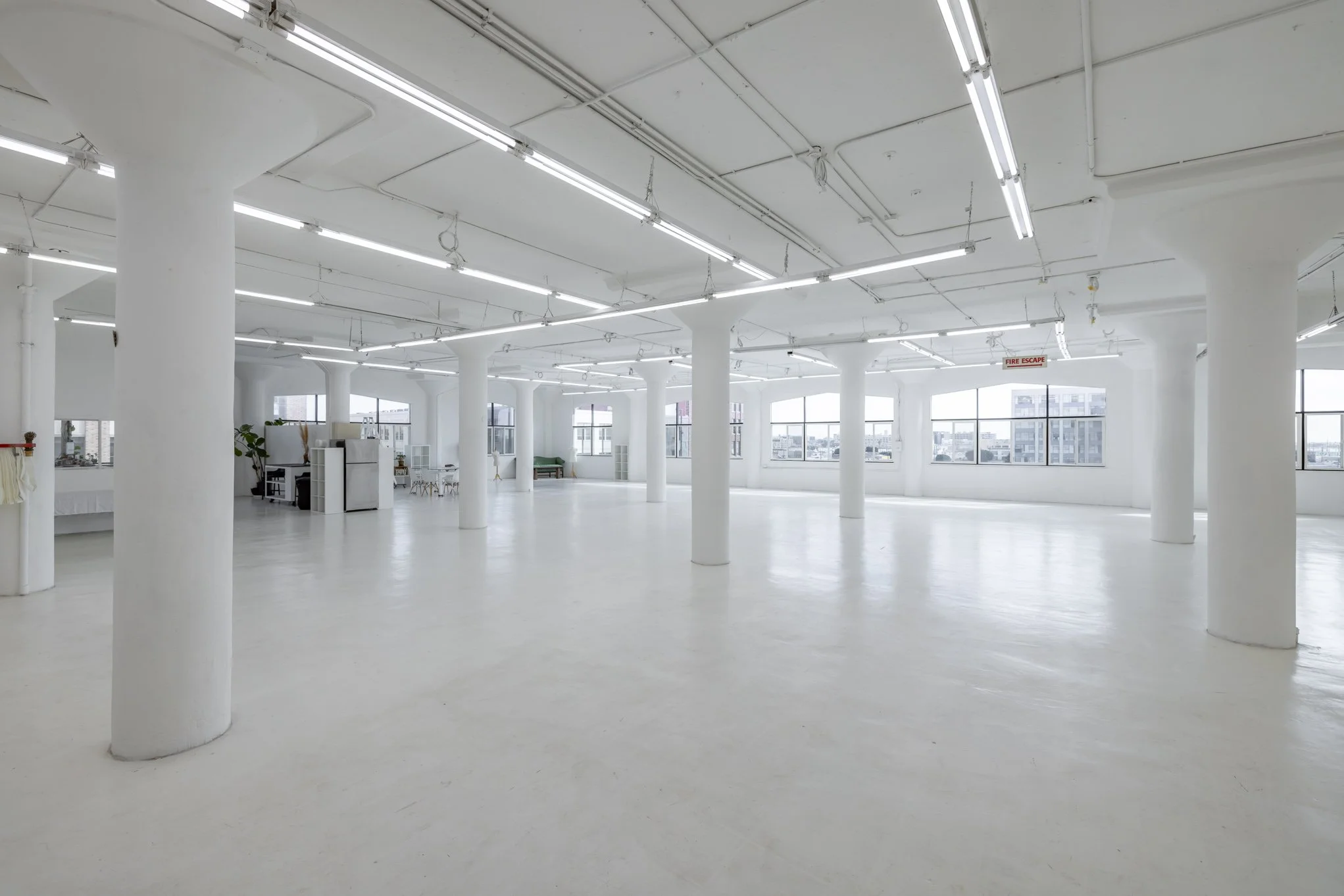A large, empty, white-walled interior space with white pillars, large windows, and fluorescent lighting