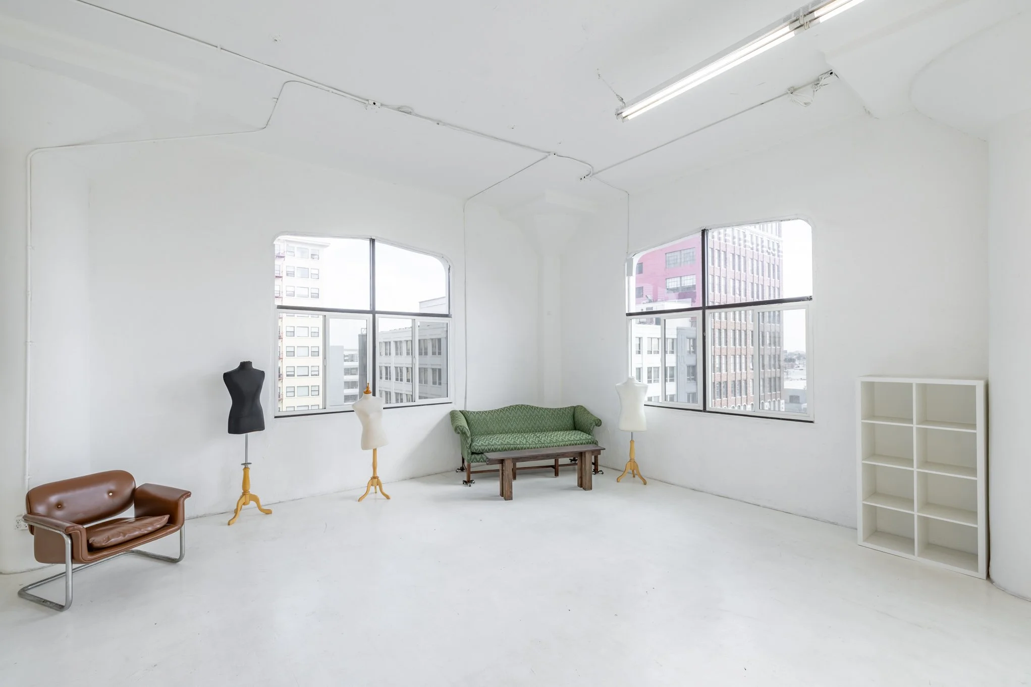 Empty white-walled room with large windows, a green couch, a small brown sofa, three mannequin torsos, and a white shelving unit, city buildings visible outside.