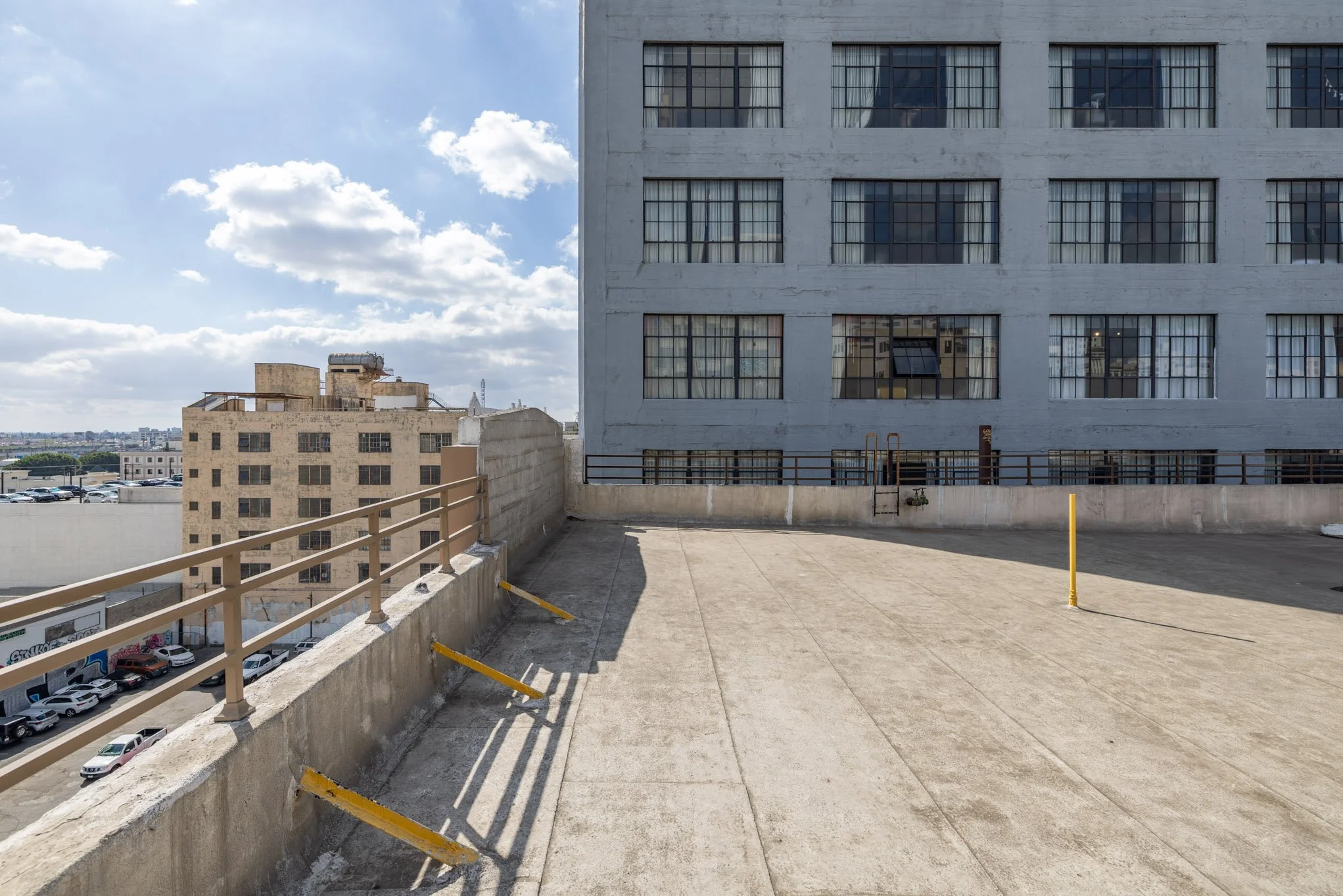 Empty rooftop parking lot surrounded by concrete walls and a metal railing, with city buildings in the background and a partly cloudy sky.