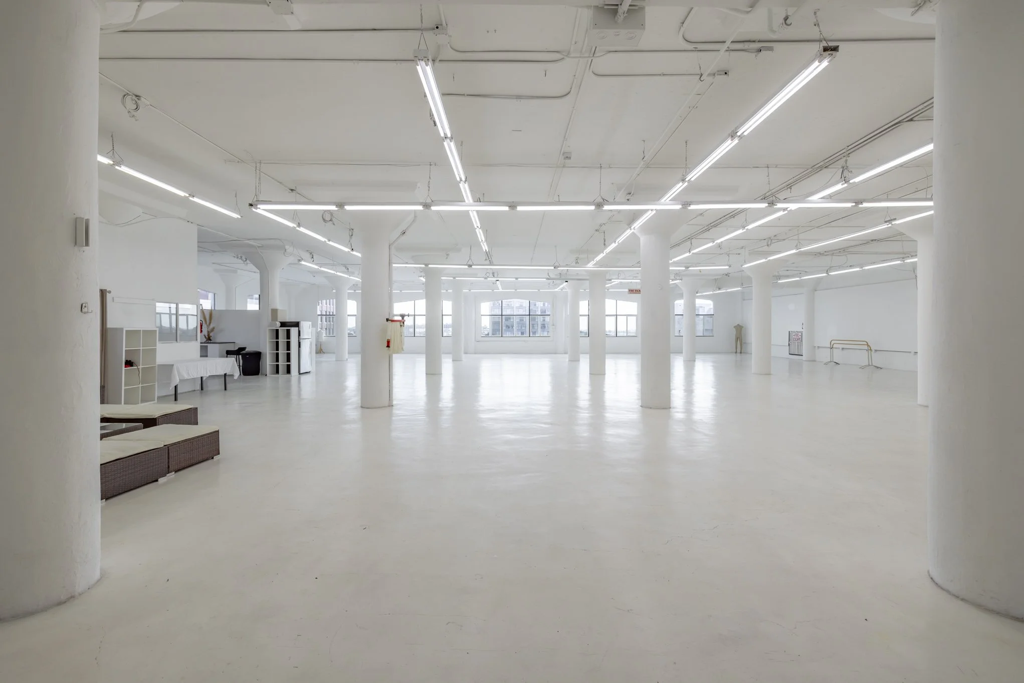 A spacious, empty, white-walled interior with large windows, white columns, and fluorescent lighting, resembling a retail or gallery space.