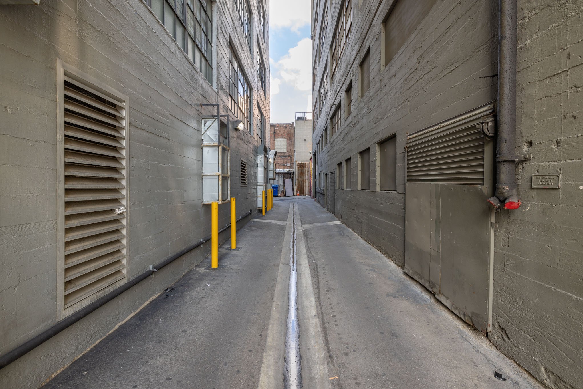 Narrow alleyway between two buildings with plain gray walls, utility vents, pipes, and yellow bollards.