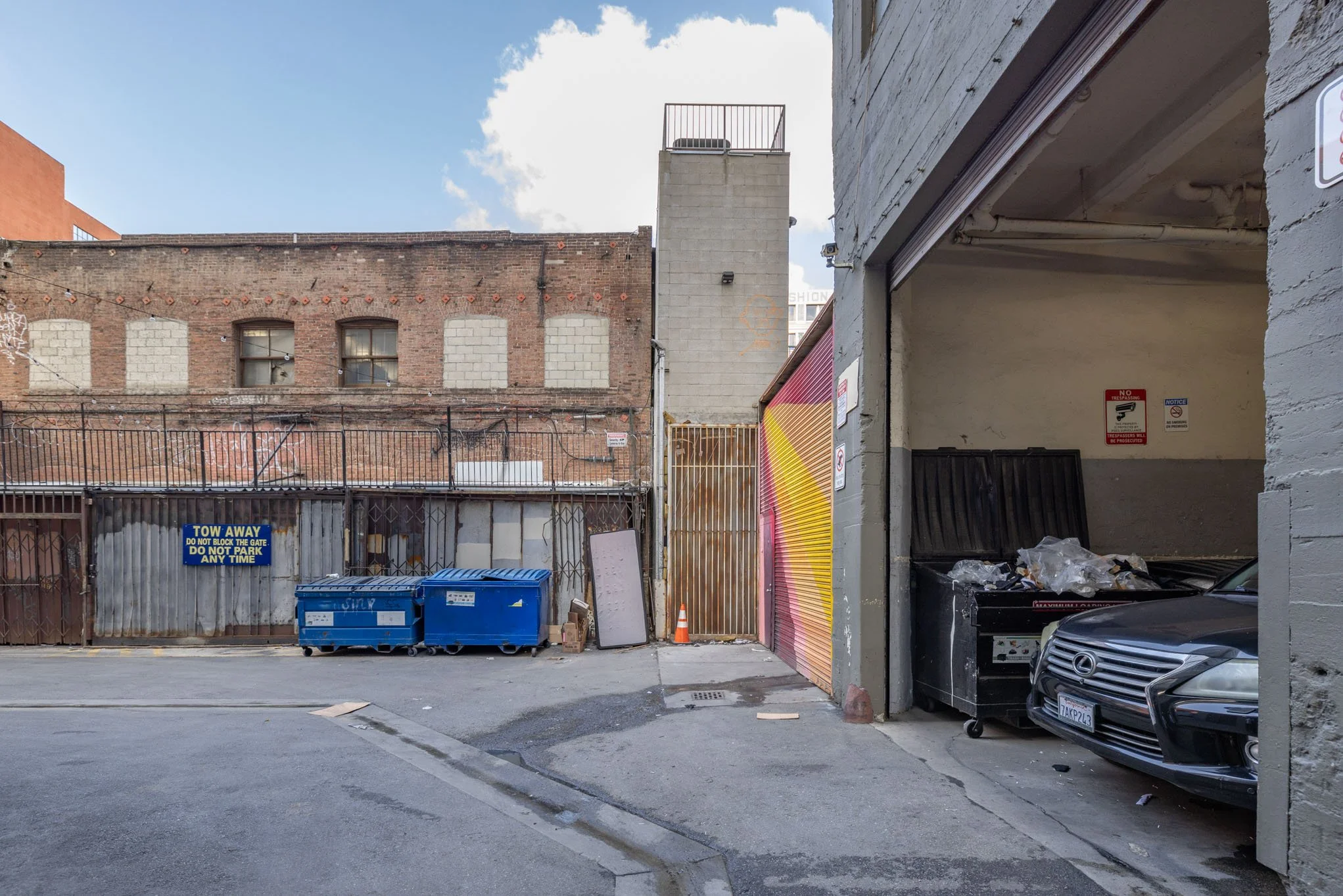 Back alley with blue dumpsters, a black Lexus car parked inside a garage, corrugated metal fences, brick and concrete buildings, and graffiti, under a partly cloudy sky.