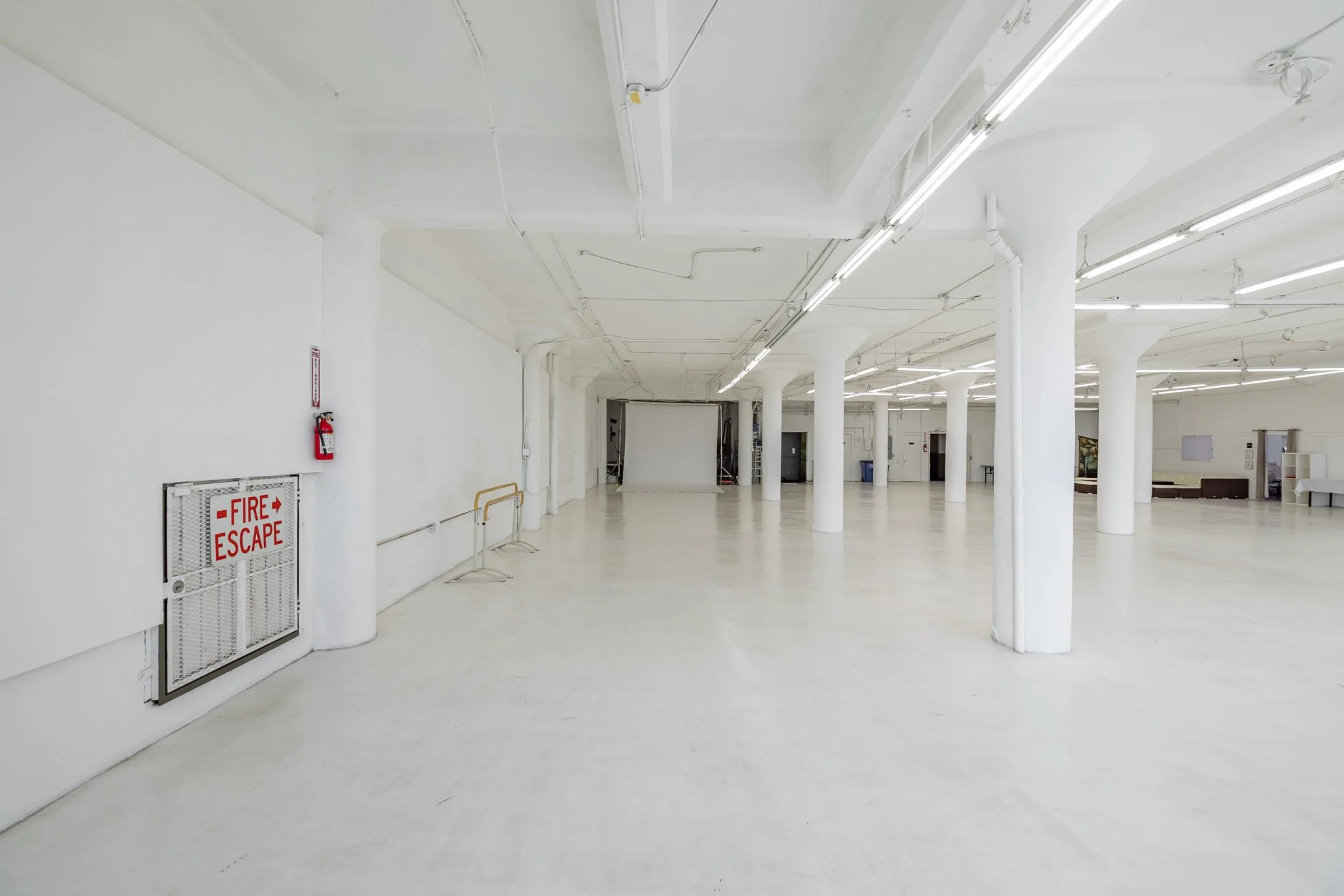 Empty white gallery with white columns, fluorescent lights, and a fire escape door on the left wall.