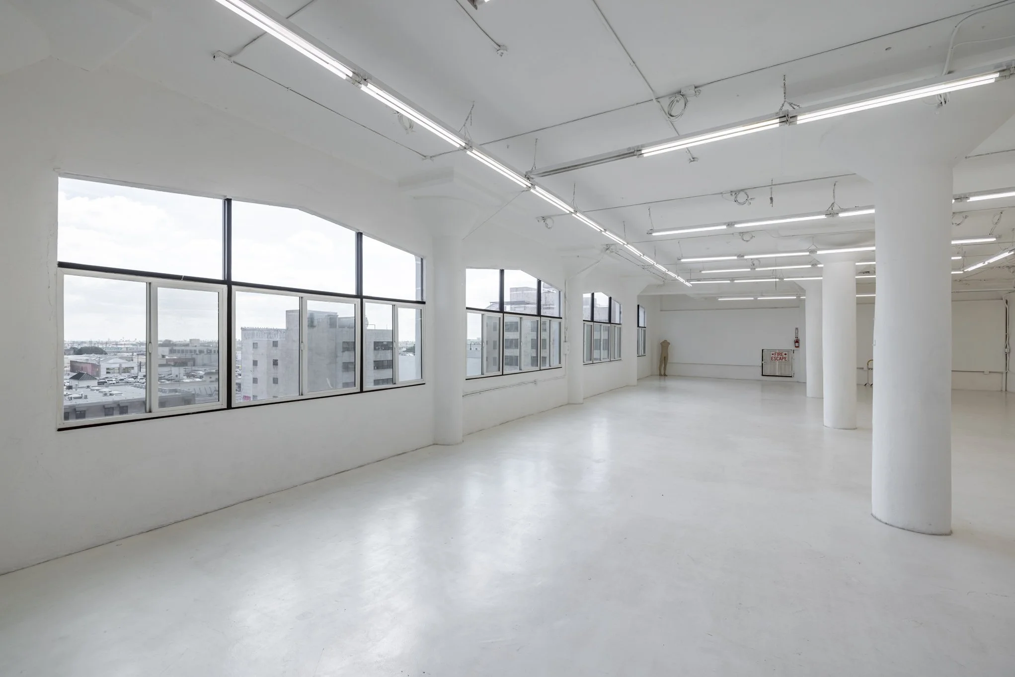 Empty white art gallery or studio with large windows, ceiling lights, and a fire escape box in the corner.