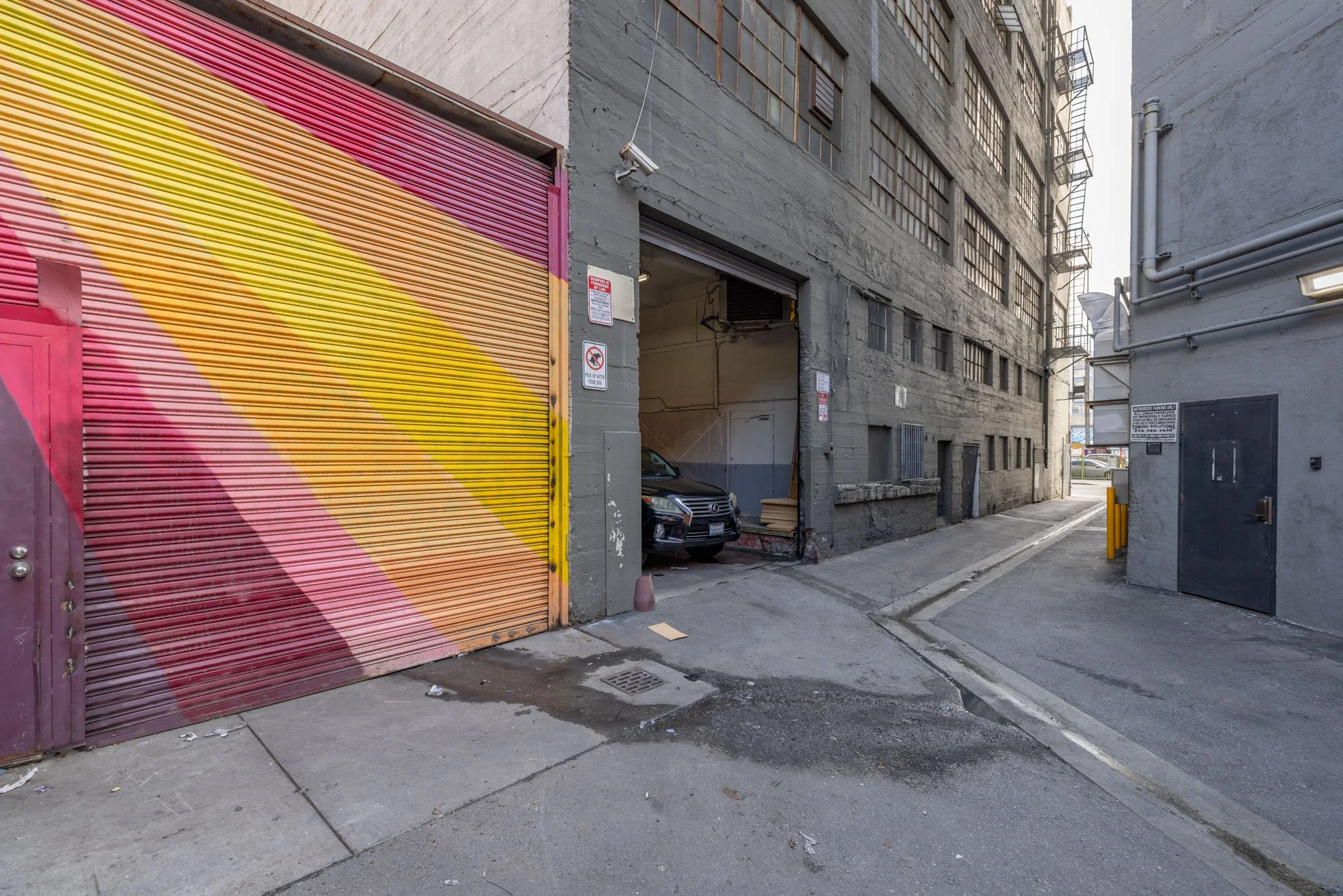 Urban alleyway with a multicolored striped metal garage door, concrete sidewalk, and a gray building with fire escapes.