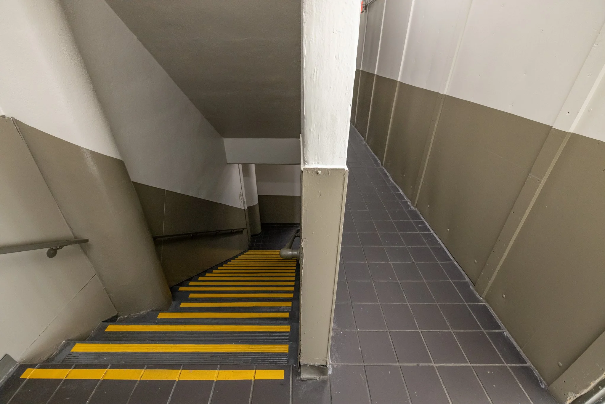 A downward view of a staircase with yellow safety striping, leading to a lower level, with a connecting corridor to the right, in a building interior.