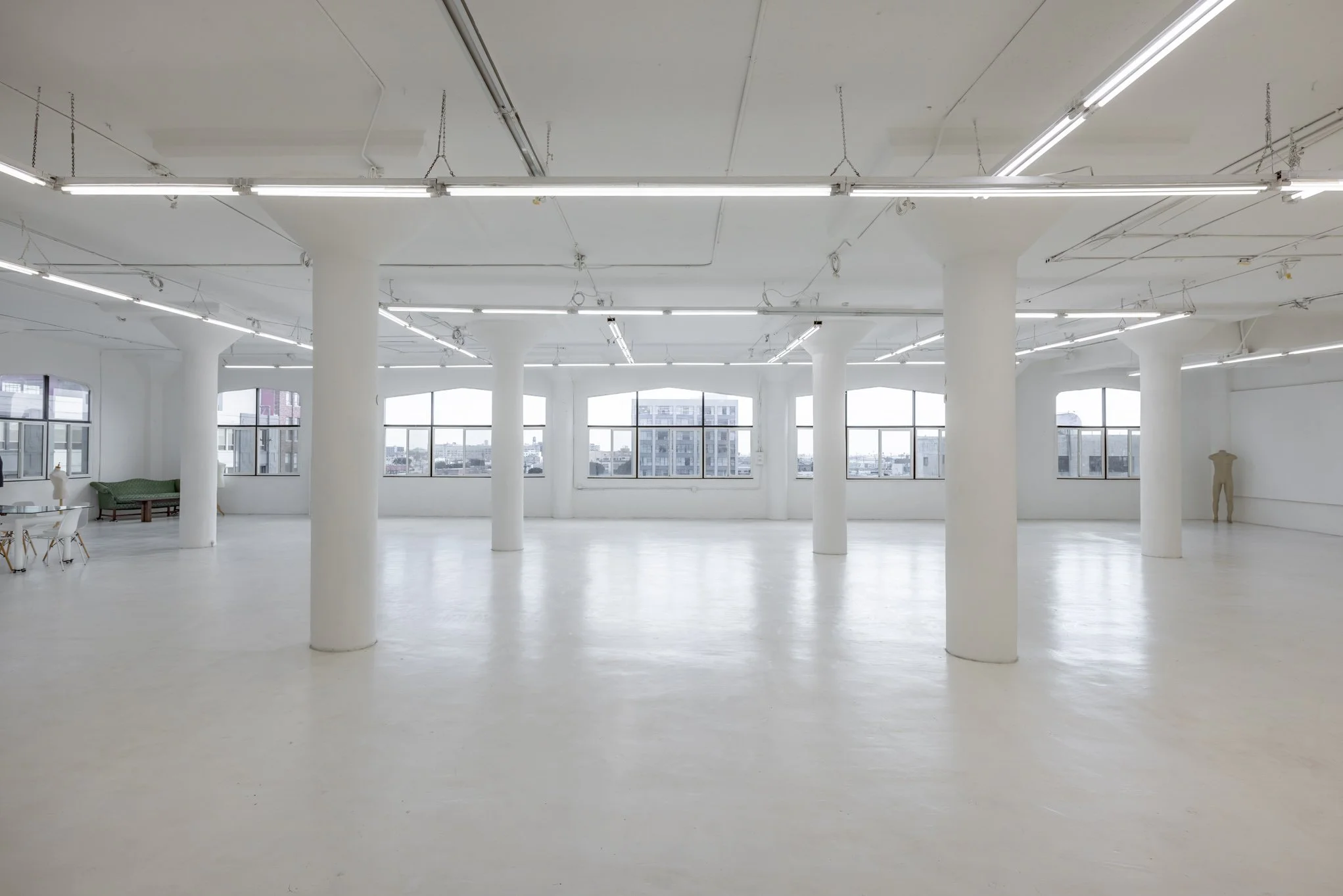 Empty, white-walled art gallery or exhibition space with large windows, white floor, and ceiling lighting.