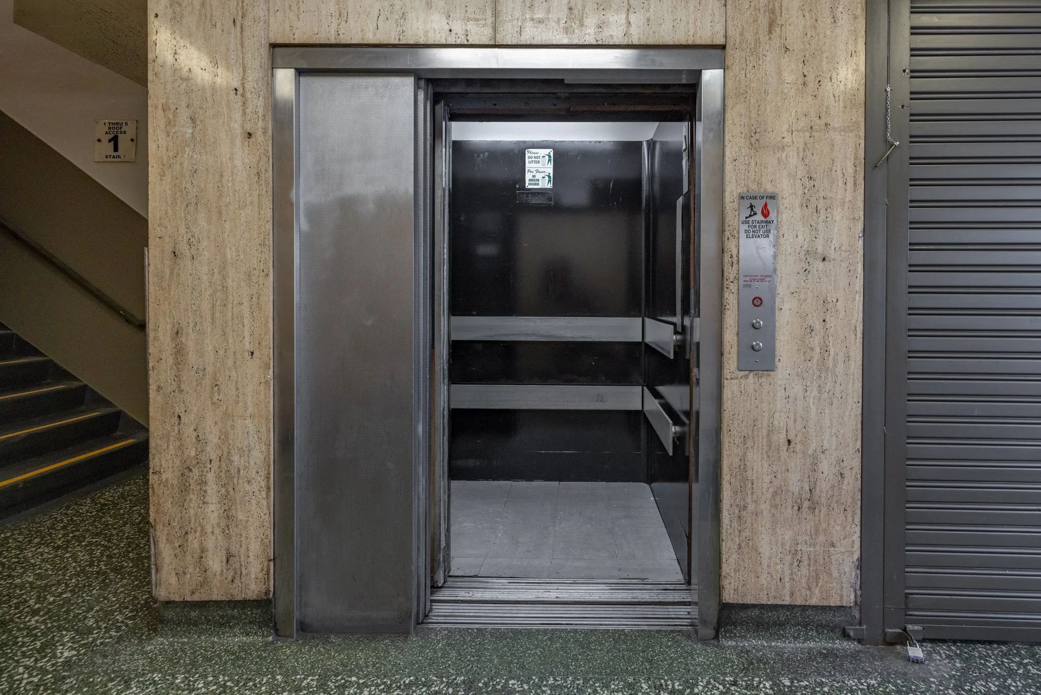 Empty elevator with black interior and stainless steel walls, situated next to a beige marble wall and a gray rolling door.