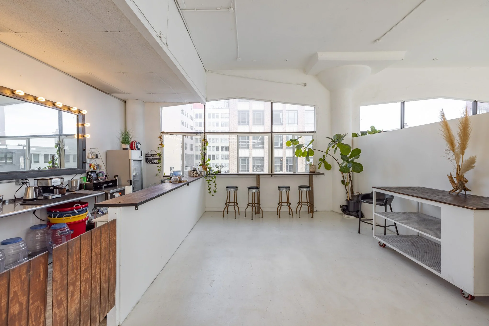 Bright, minimalist indoor space with large windows, potted plants, bar stools, and a long counter with coffee-making equipment.