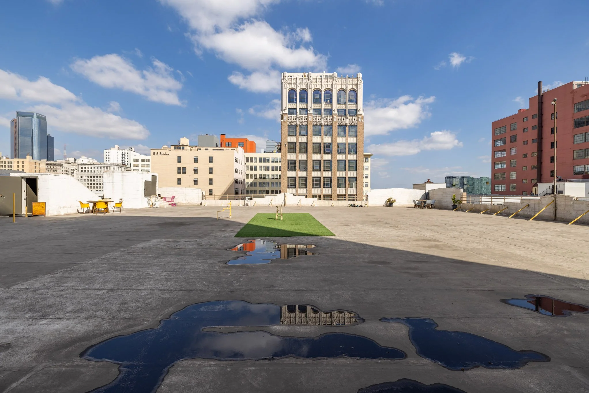 Empty rooftop parking lot with puddles of water, a small patch of green grass, surrounded by city buildings under blue sky with clouds.