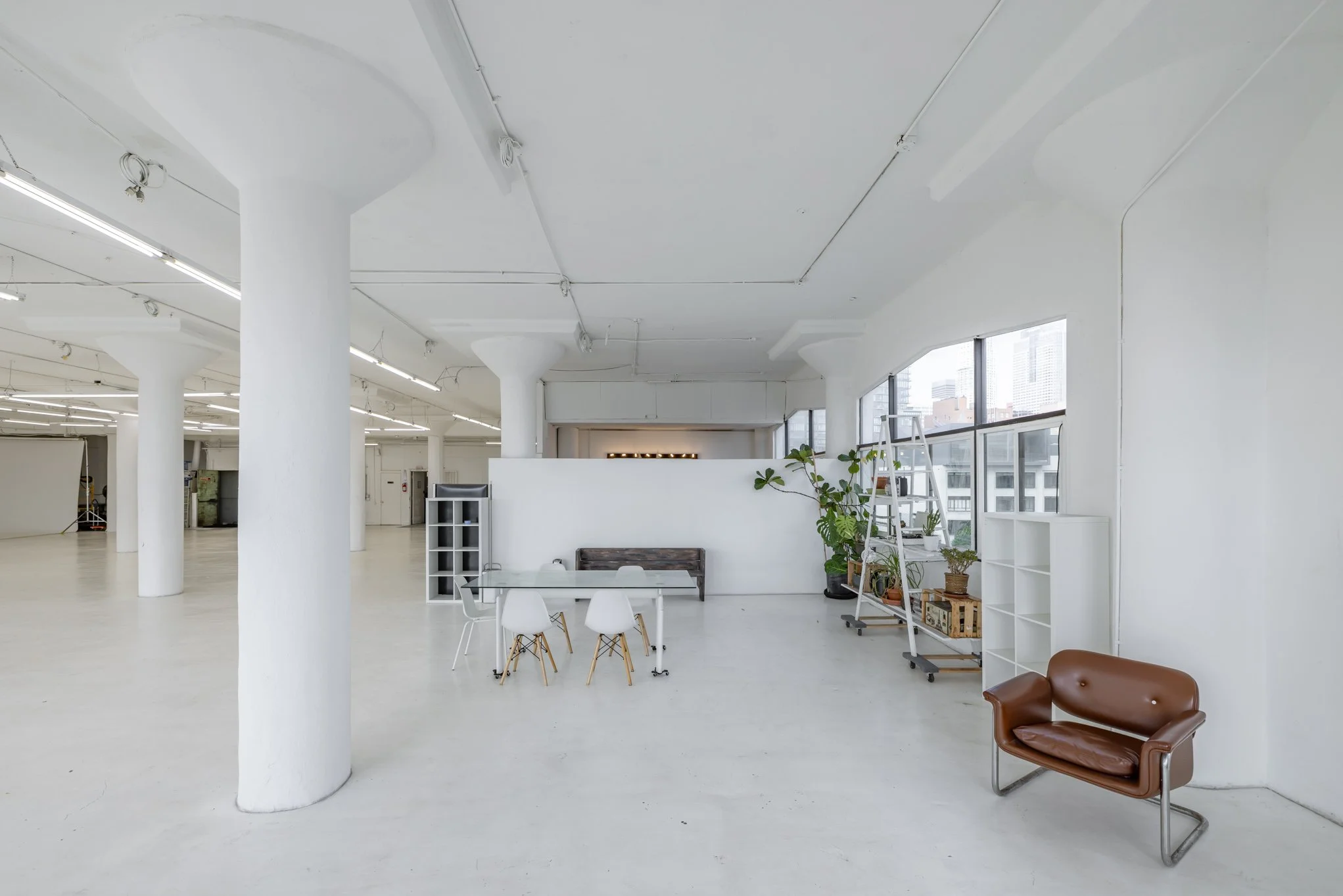 An empty, modern, white-walled interior space with large windows, a brown leather chair, a white shelving unit, a ladder with potted plants, and a glass table with white chairs.