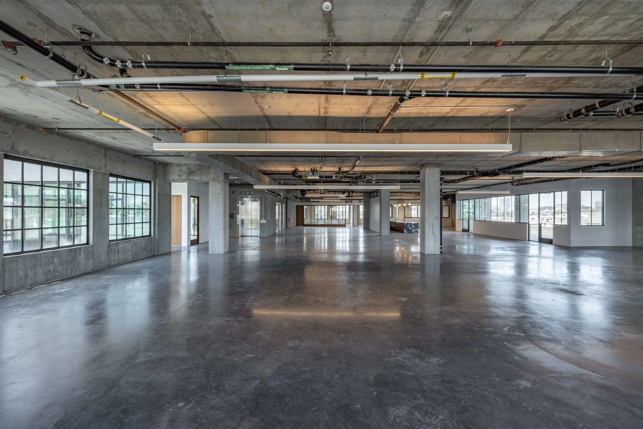 Empty commercial space with concrete floors, exposed ceiling pipes, large windows, and natural light.