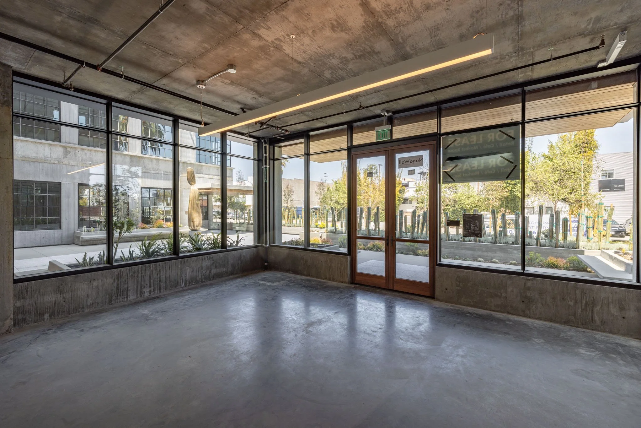 Empty commercial space with large glass windows, concrete floors, and visible industrial ceiling fixtures, overlooking a landscaped outdoor area.