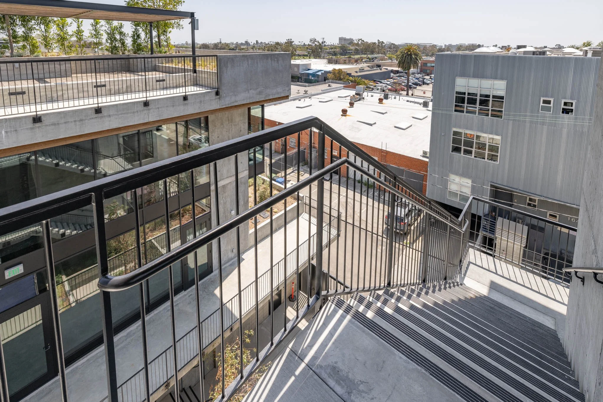 View from an outdoor staircase on a balcony overlooking a cityscape with modern commercial and residential buildings, parking lot, and trees in the background.