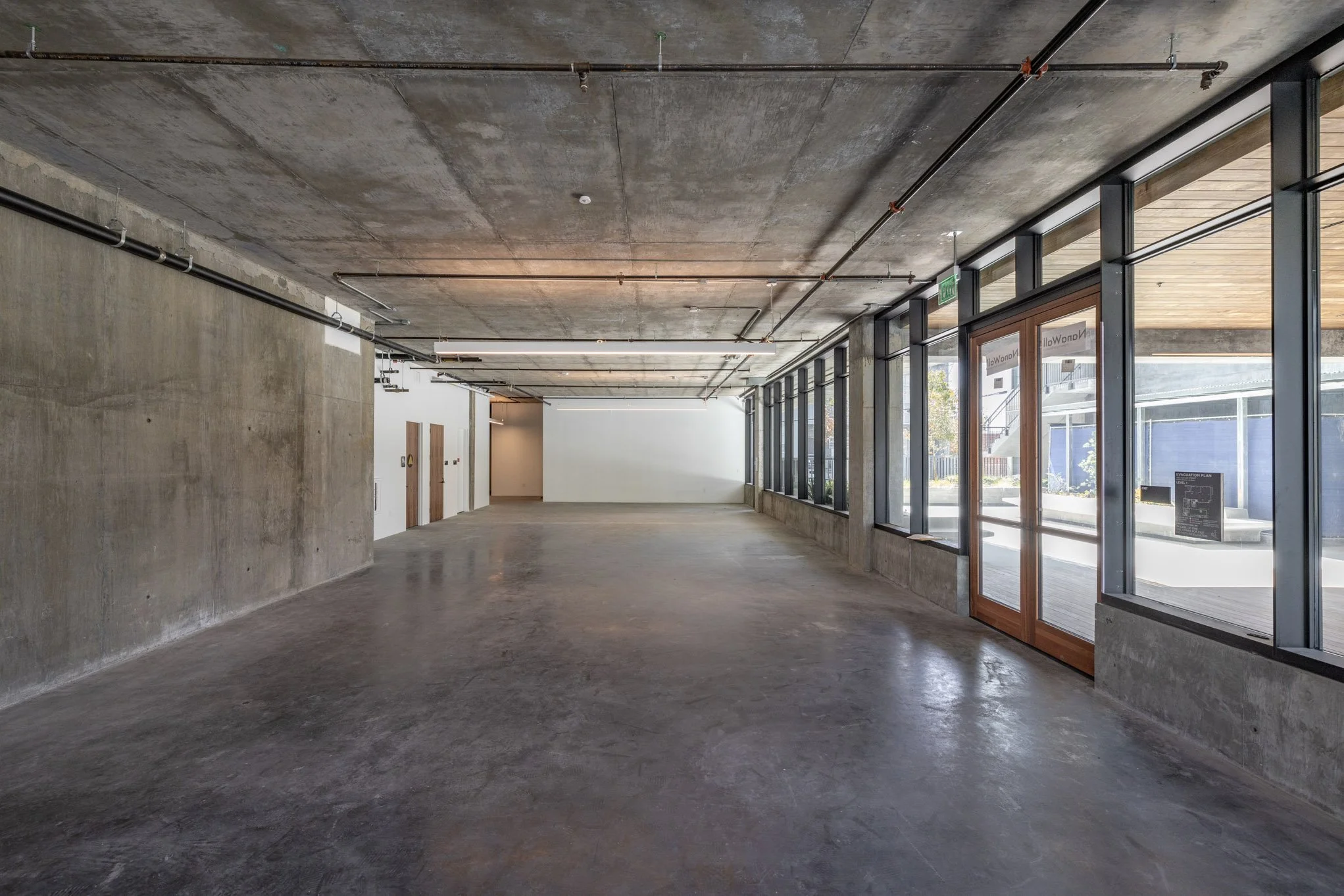 Empty modern commercial space with large glass windows, concrete floors, and exposed ceiling with visible pipes.