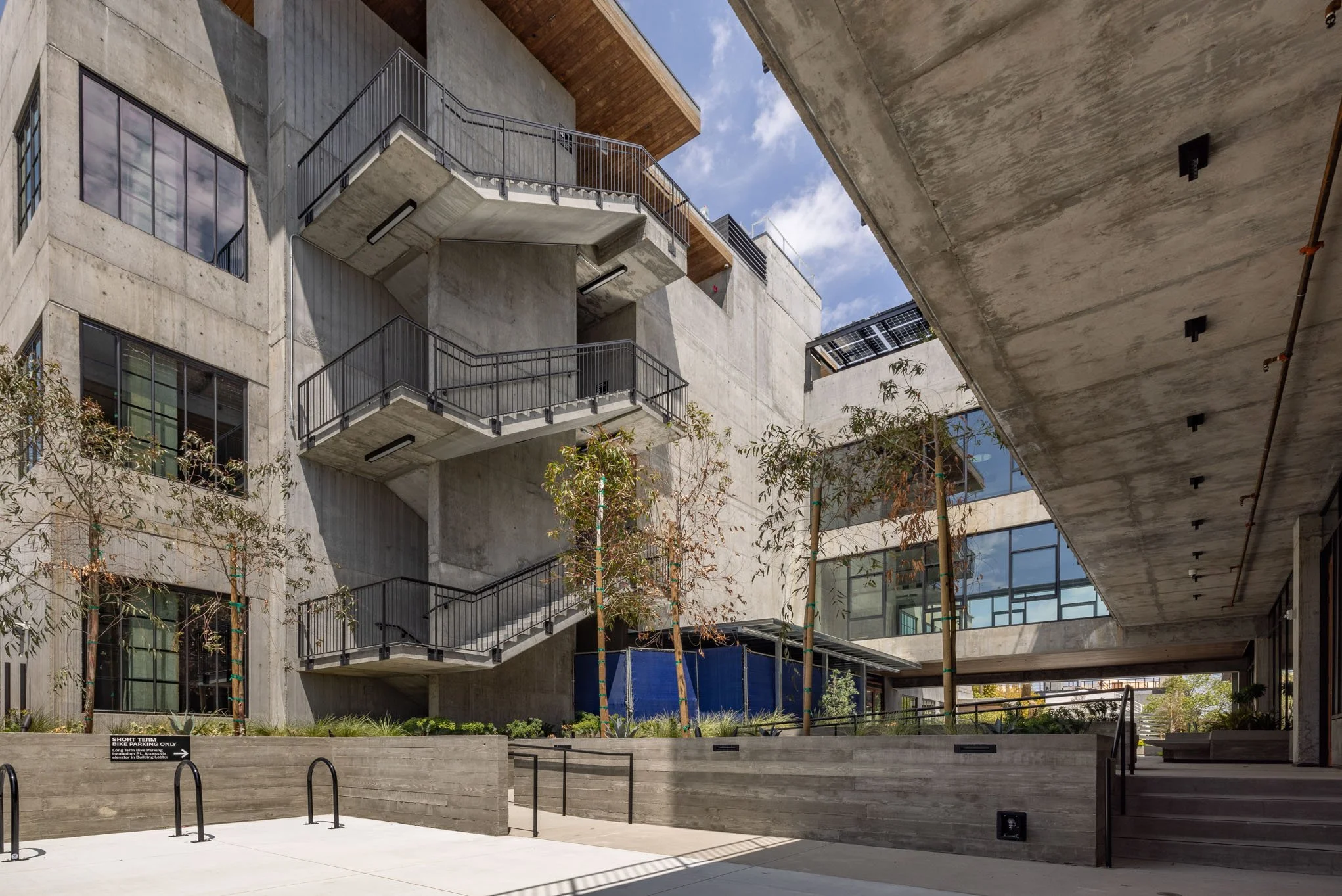 Modern multi-story concrete building with exterior stairs, large windows, and landscaped trees and bushes in an outdoor courtyard.