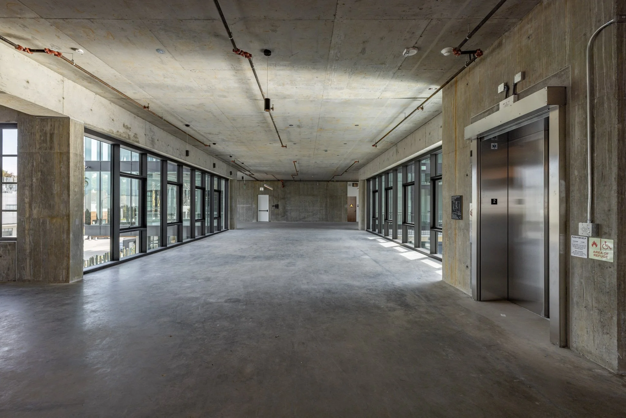 Empty multi-level parking garage with concrete floors, walls, and ceiling, large windows on both sides, an elevator on the right, and exposed pipes and wiring on the ceiling.